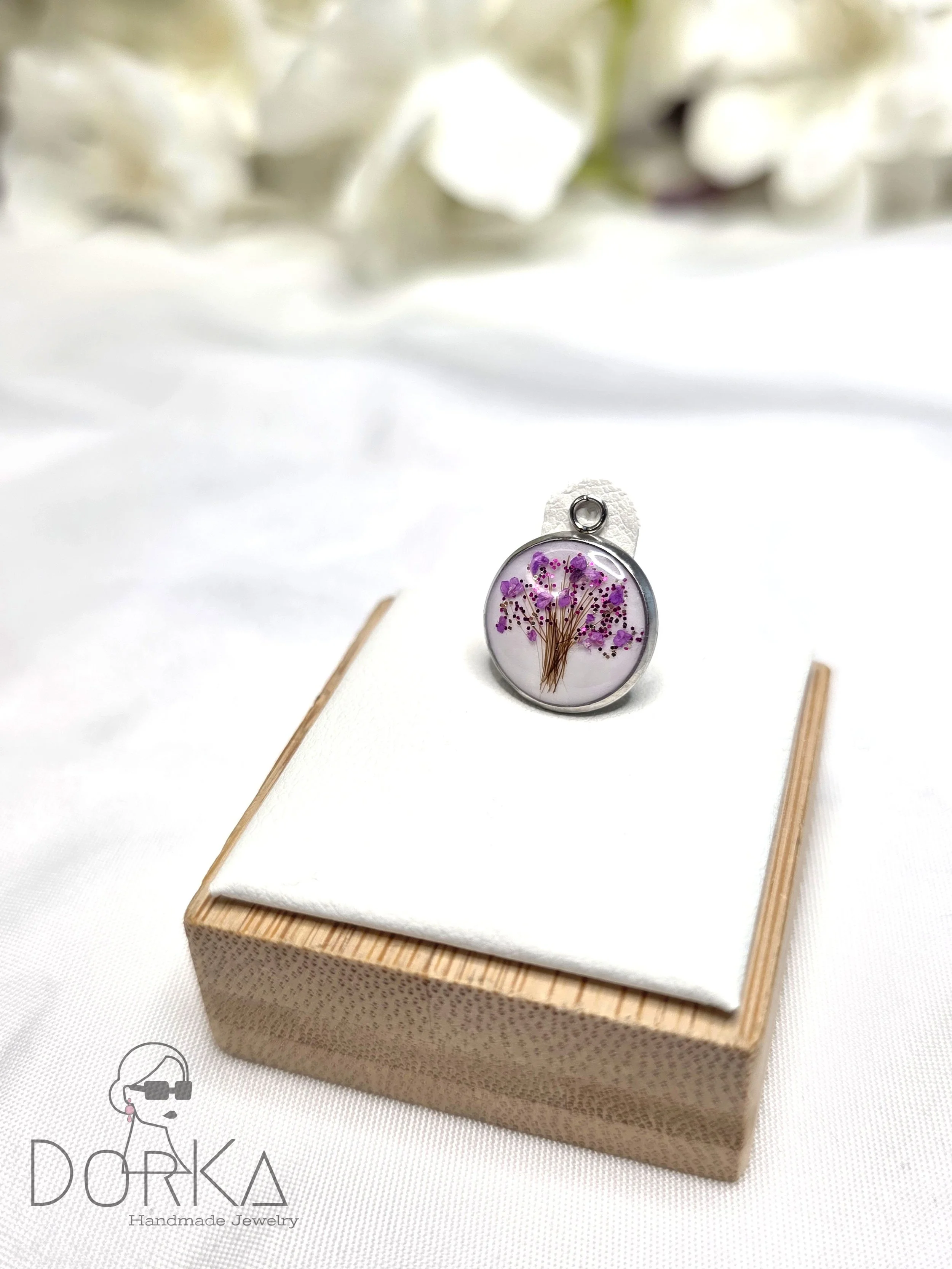 Ein Ringschmuckstück mit eingegossenem getrocknetem Blumenarrangement, das auf einer weißen Stoffunterlage auf einem kleinen Holzsockel liegt.