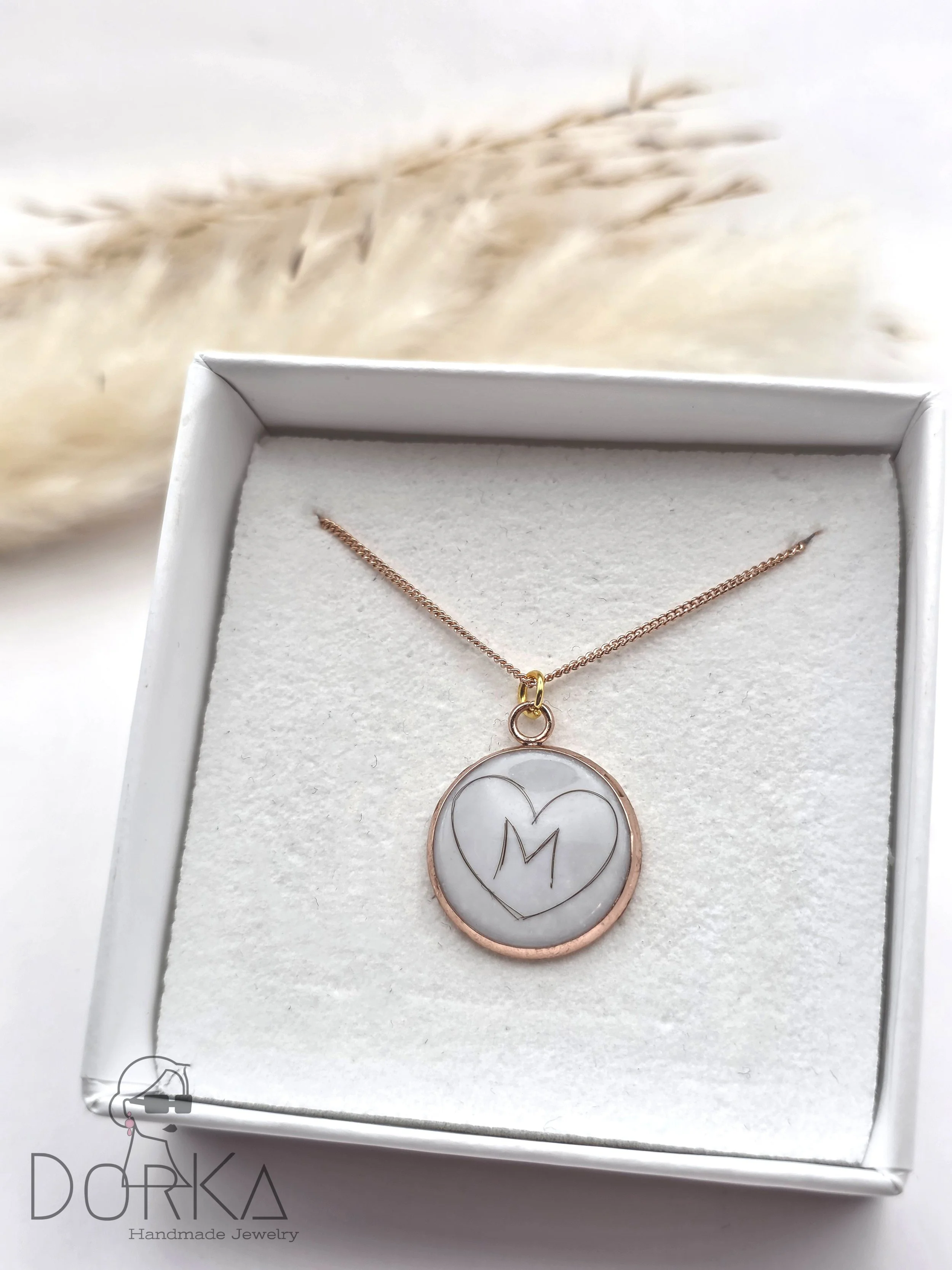 Ein handgefertigtes Schmuckstück, eine Halskette mit einem runden Anhänger, der ein Herz mit einem Buchstaben M darin zeigt, befindet sich in einer weißen Schmuckschachtel.