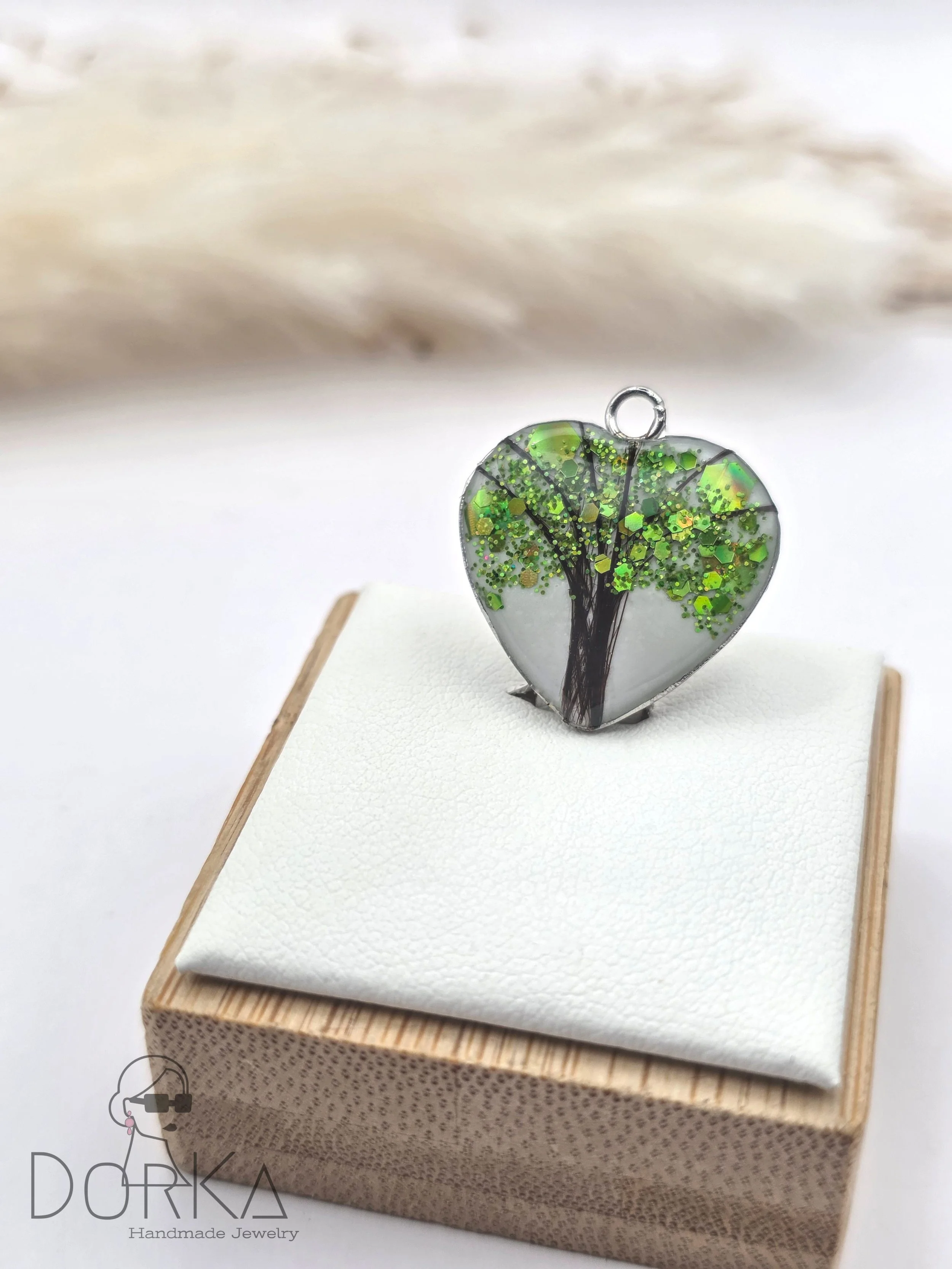 Herzförmiger Anhänger mit grünen Glitzerregentröpfchen und Baumdesign, auf weißem Leder auf einem Holz- und Stoffkissen, von Dorka Handmade Jewelry.