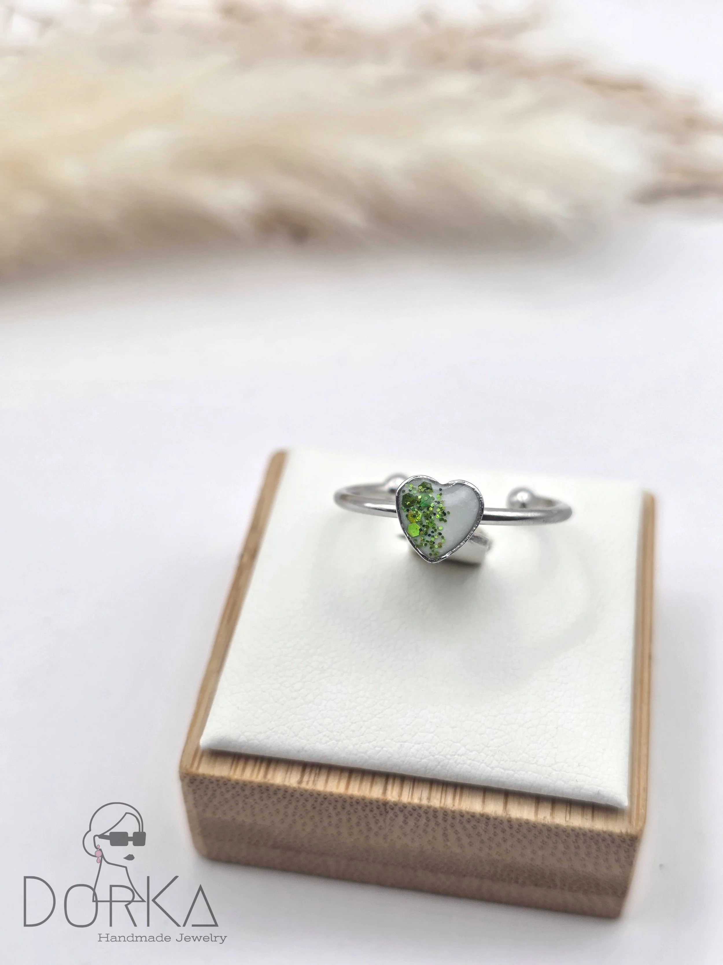 Silberner Ring mit Herzform und grünen Glitzersteinen auf einem weißen Schmuckkissen in einer Holzkiste.