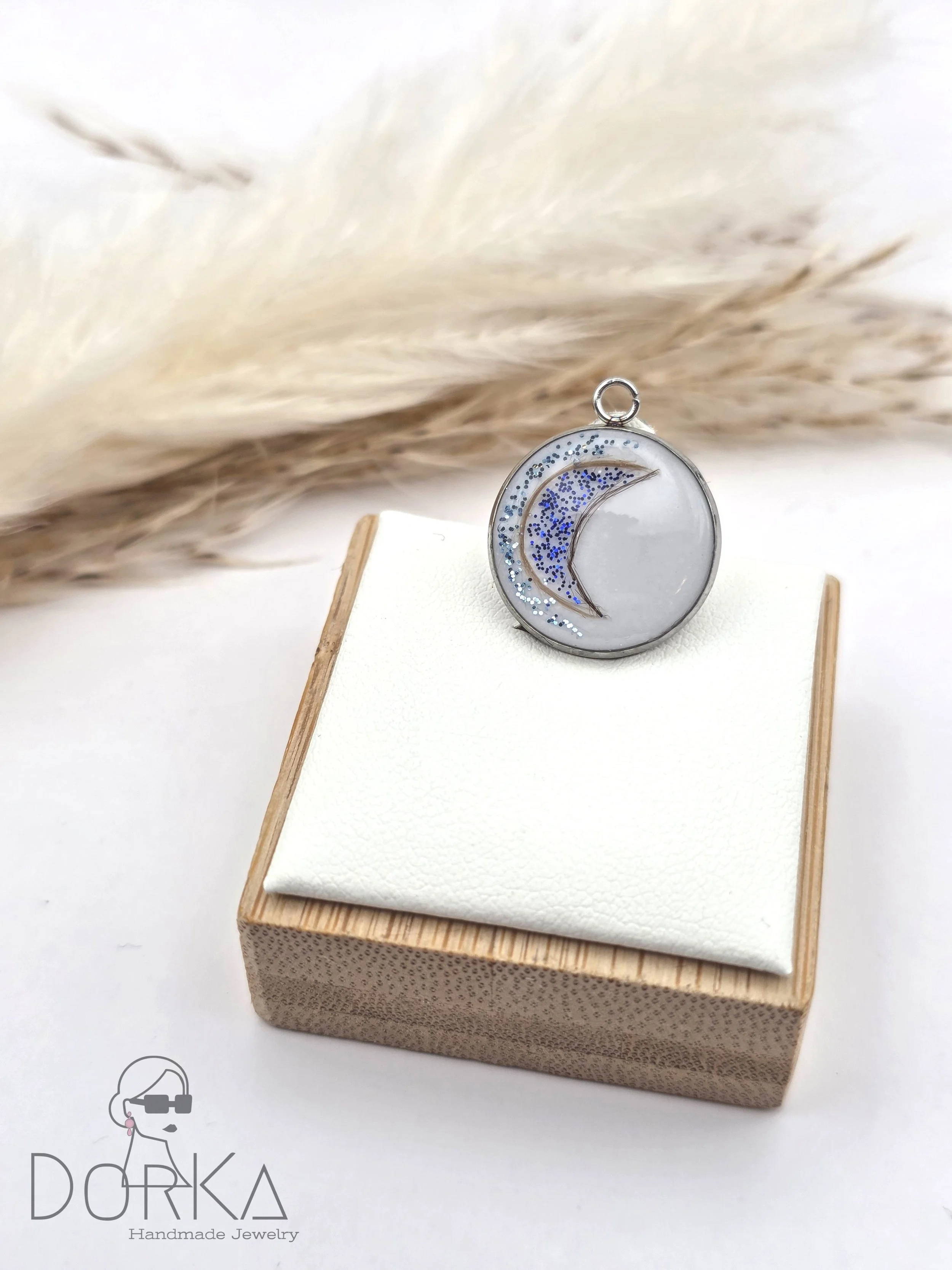 Hanging Anhänger mit Mond-Design, blauen Glitzer und Silber-Details auf weißem Hintergrund, auf eine weiße Hartschaumgummisplatte auf einem Holzkästchen gestellt, mit einem pampasartigen Grashalm im Hintergrund.