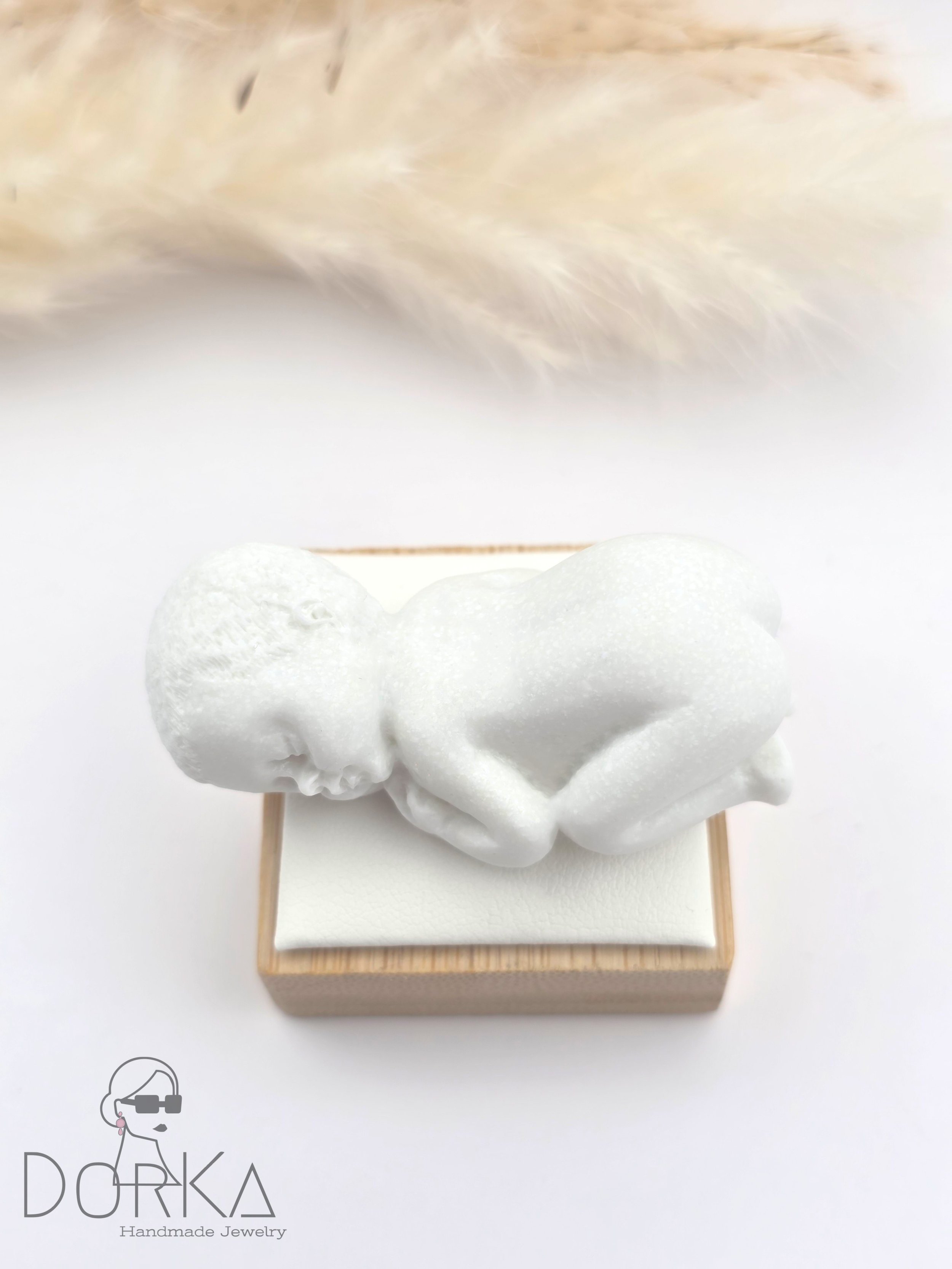 Weiße Skulptur eines schlafenden Babys auf einem kleinen Holzsockel, mit einer oberen Spiegelung eines Federkopfes im Hintergrund.