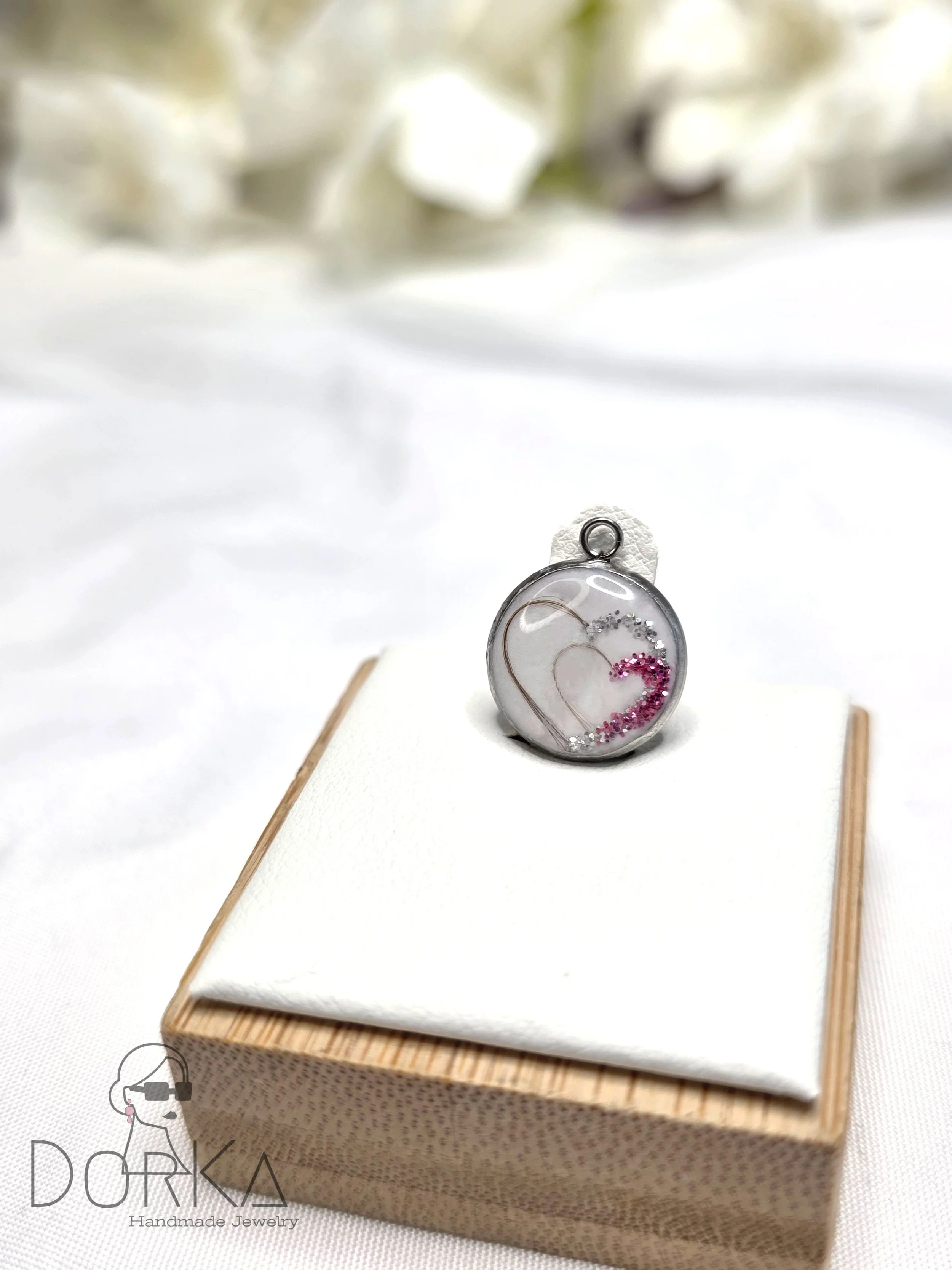 Runder Anhänger mit Herzdesign und glitzernden Dekorationen in pink und silber, auf einem weißen Kissen auf einem Holzrahmen präsentiert.