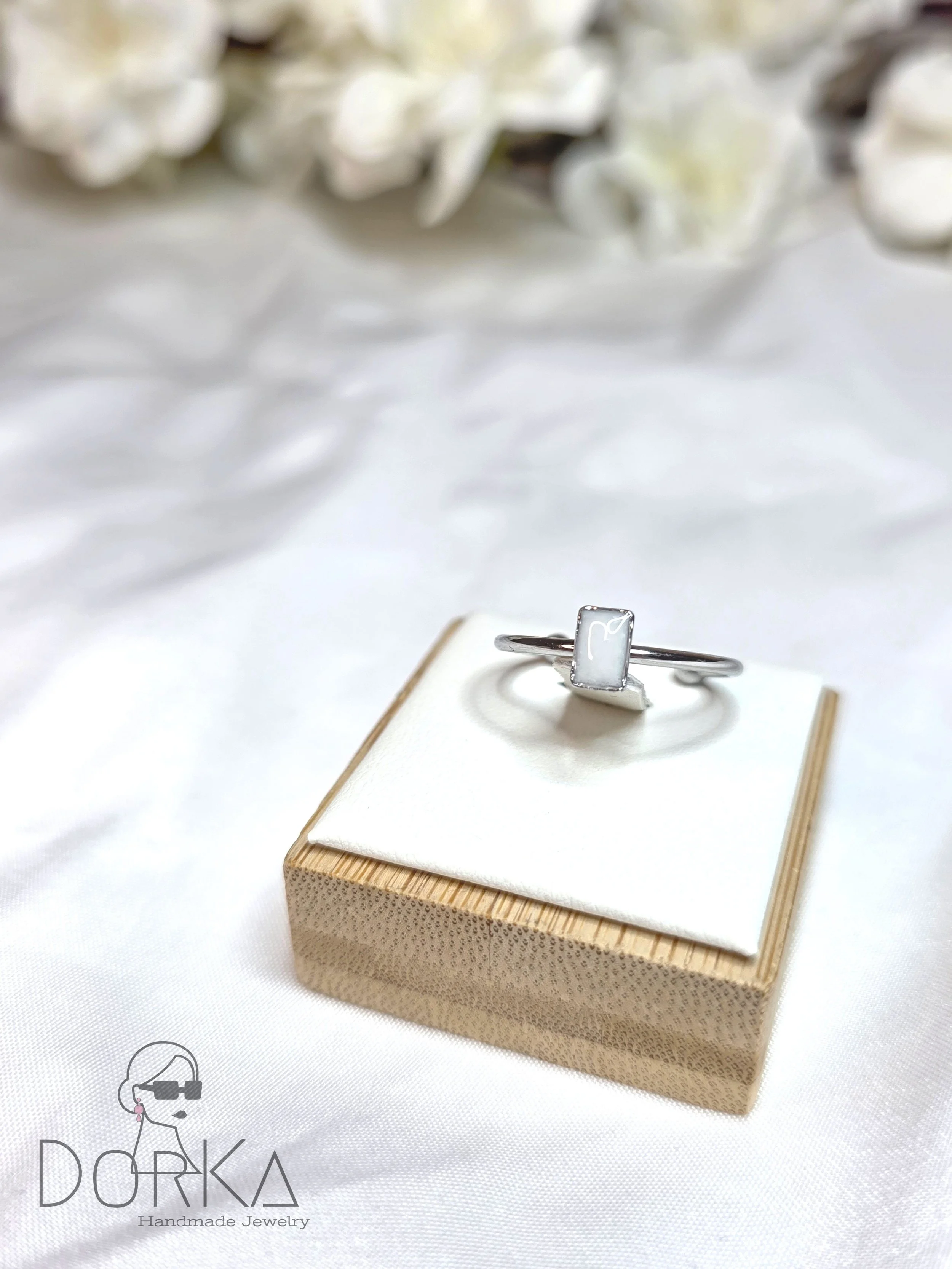 Silberring mit rechteckigem weißen Stein auf weißem Kissen in einer Papierbox, Hintergrund mit weißen Blumen.