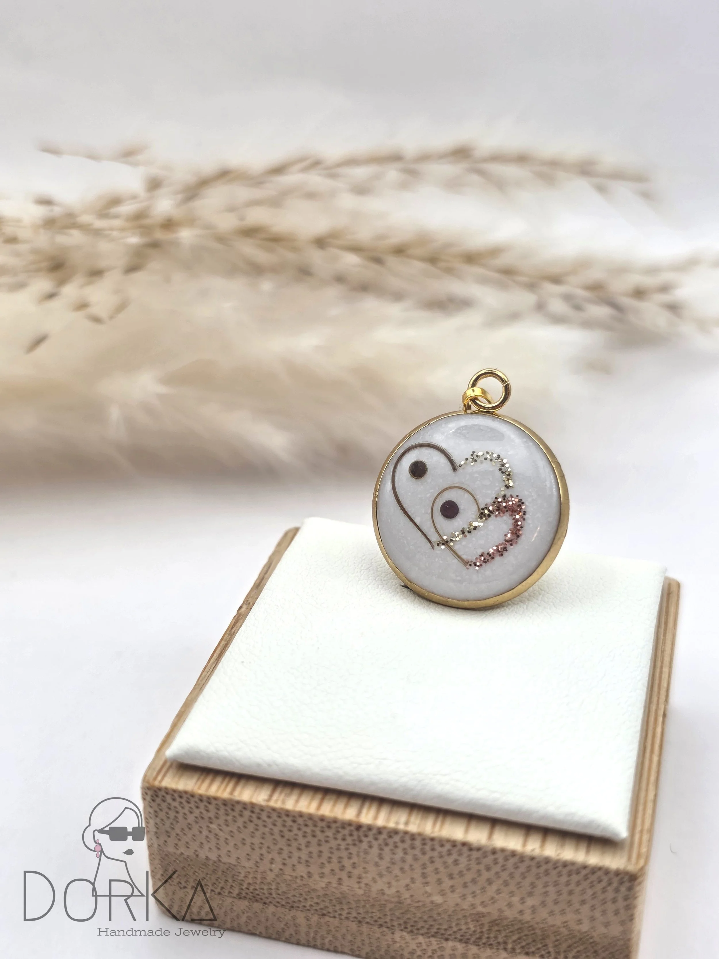 Runder Anhänger mit Herz- und Kreisdesign, goldfarbenen Rahmen, auf weißem Hintergrund, auf einem weißen Stoffkissen in einer Holzbox, DORI Handmade Jewelry Logo im unteren linken Eck.