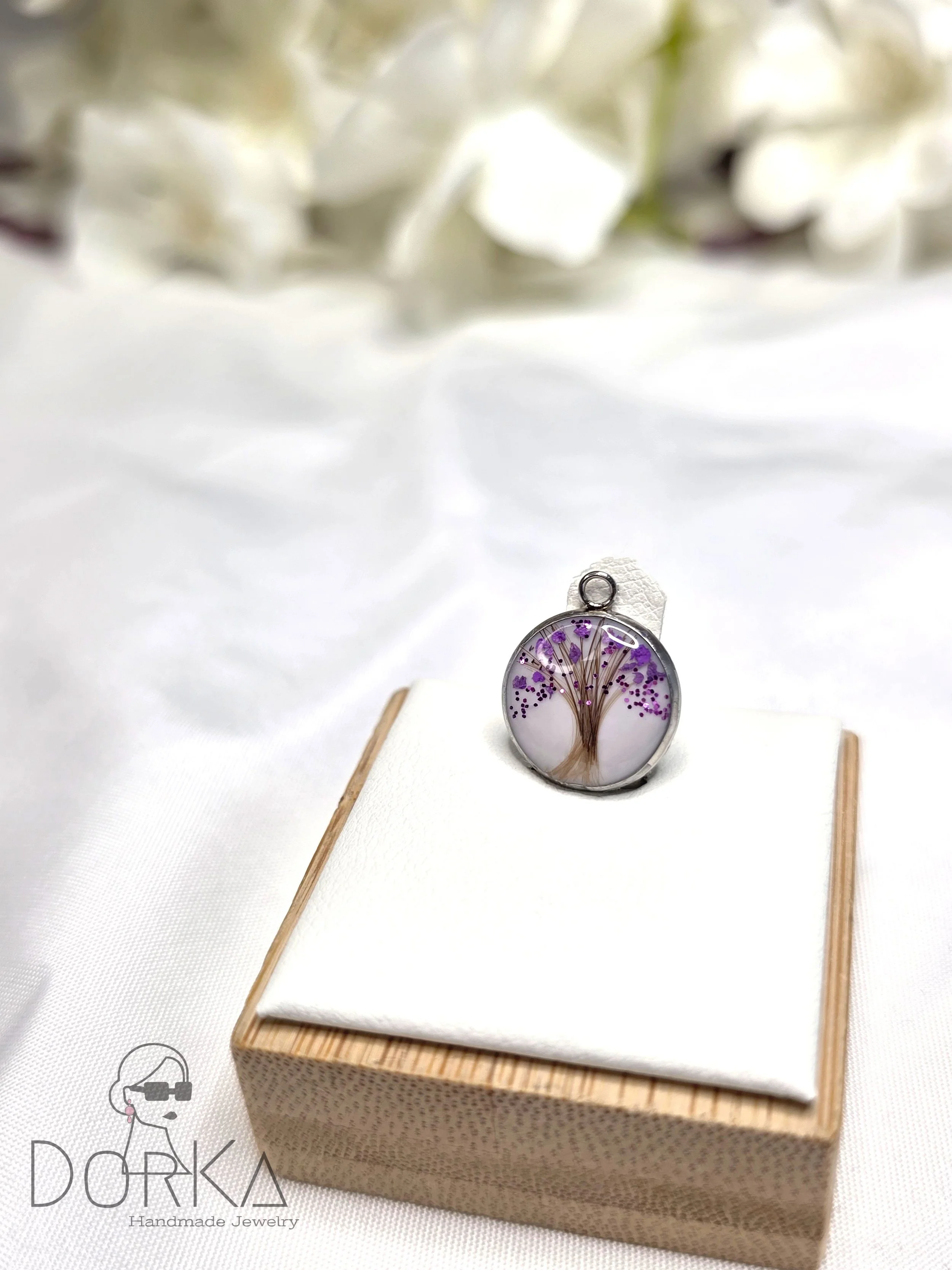 Runder Anhänger mit Baumdesign und lilafarbenen Blättern, auf weißem Tuch und Holzschachtel, mit weißen Blumen im Hintergrund.