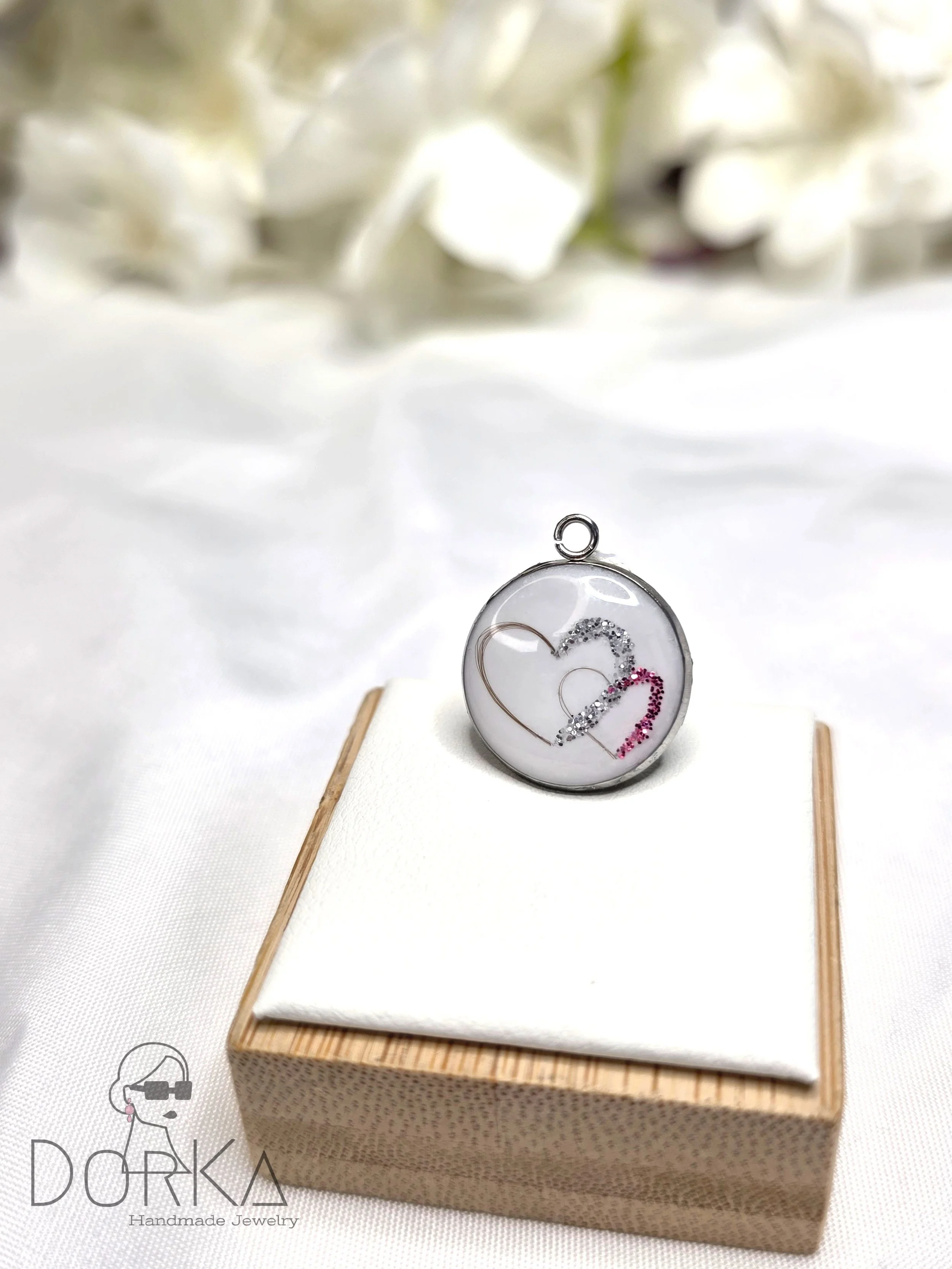 Halskette mit zwei Herzen, verziert mit weißen, schwarzen und pinken Schleifen, liegt auf einer weißen Unterlage in einer Holzkiste, im Hintergrund weiße Blumen.