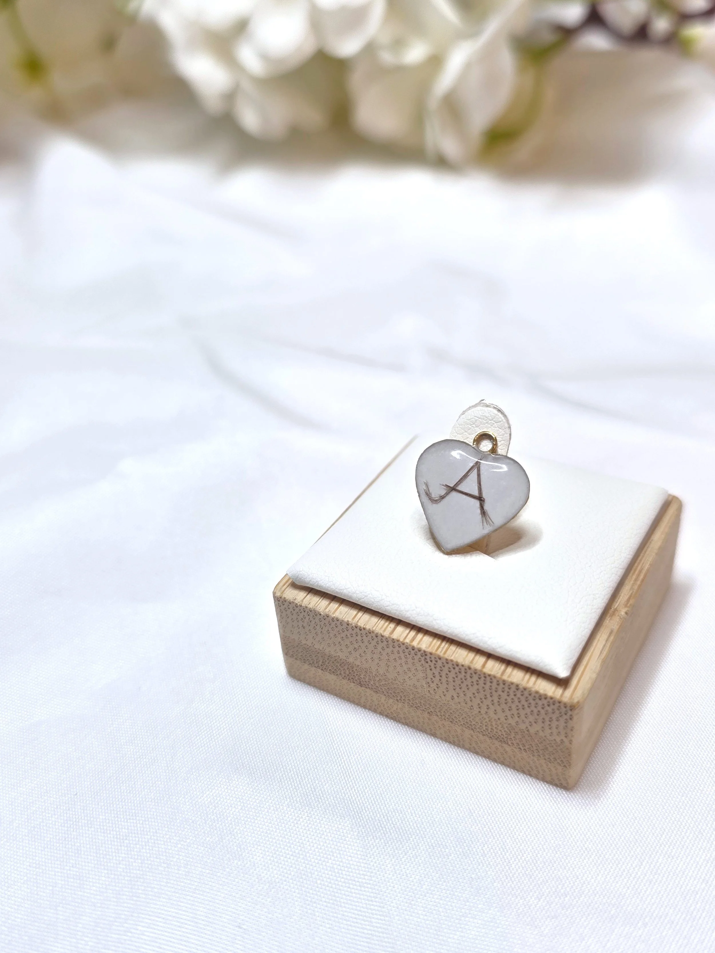 Eine kleine Schmuckschatulle mit einem Herzanhänger in Silberfarbe mit eingraviertem Buchstaben A auf einem weißen Kissen, im Hintergrund weiße Blumen.