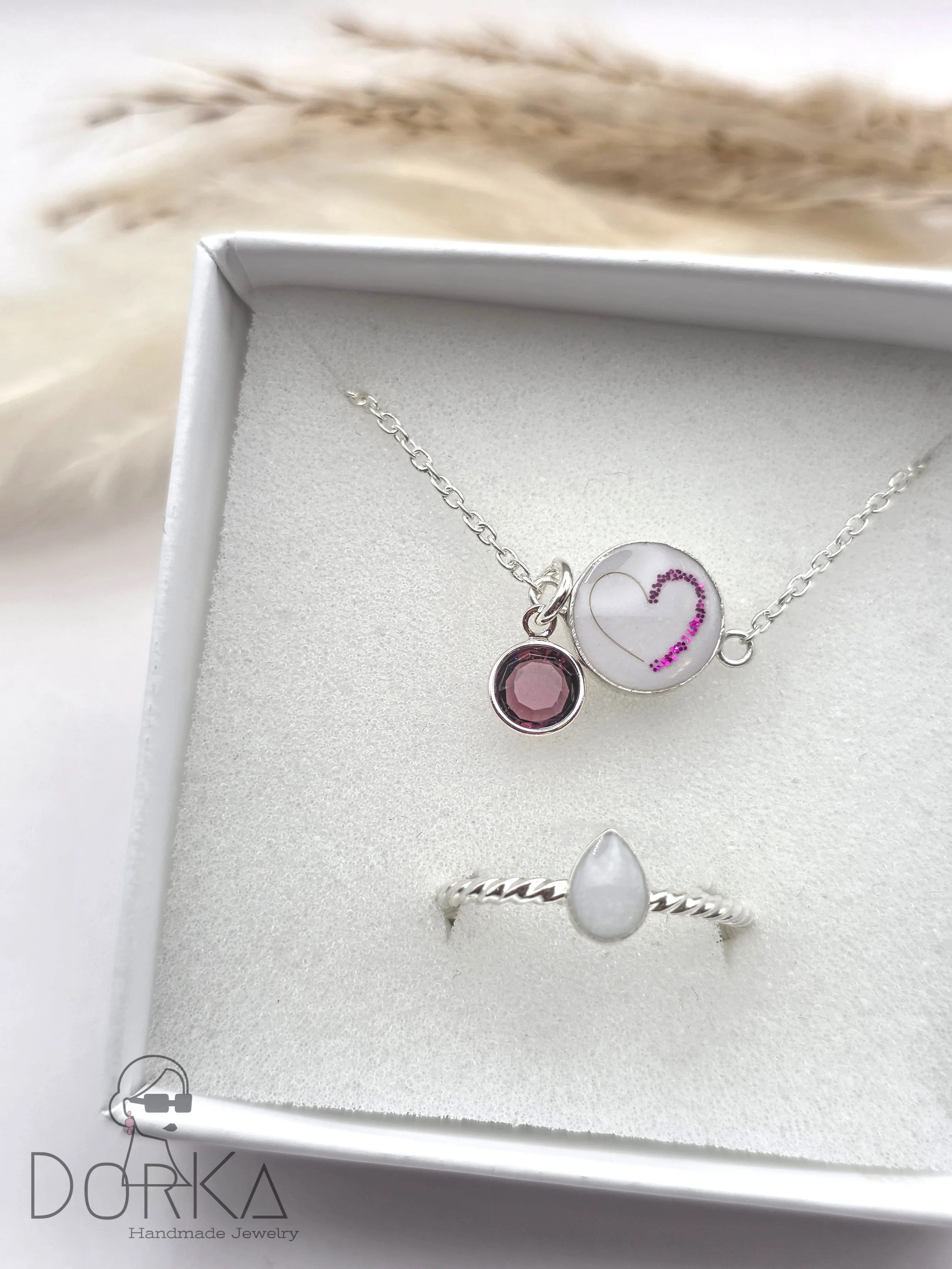 Silberne Kette mit zwei Anhängern, einem weißen mit lila Fragezeichen, und einem pinken Edelstein, sowie ein Silber-Ring mit einer weißen Perle in einem weiße Schmuckschatulle