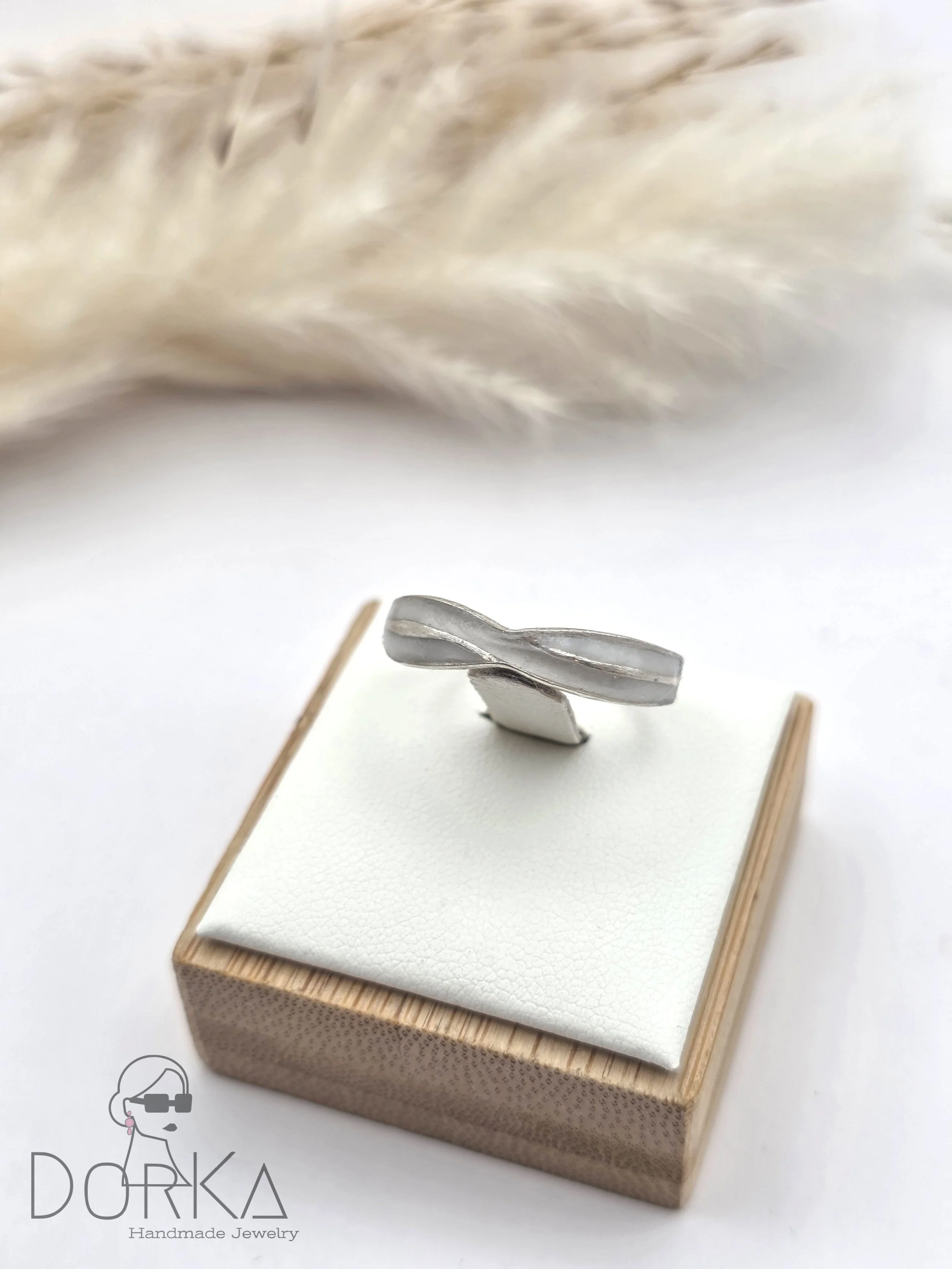 Ein silberner handgefertigter Schmuckring auf einem weißen Samtuntergrund auf einer kleinen Holzkiste, im Hintergrund ein vages Fell. Unter dem Bild befindet sich ein Logo mit der Aufschrift 'Dorka Handmade Jewelry'.