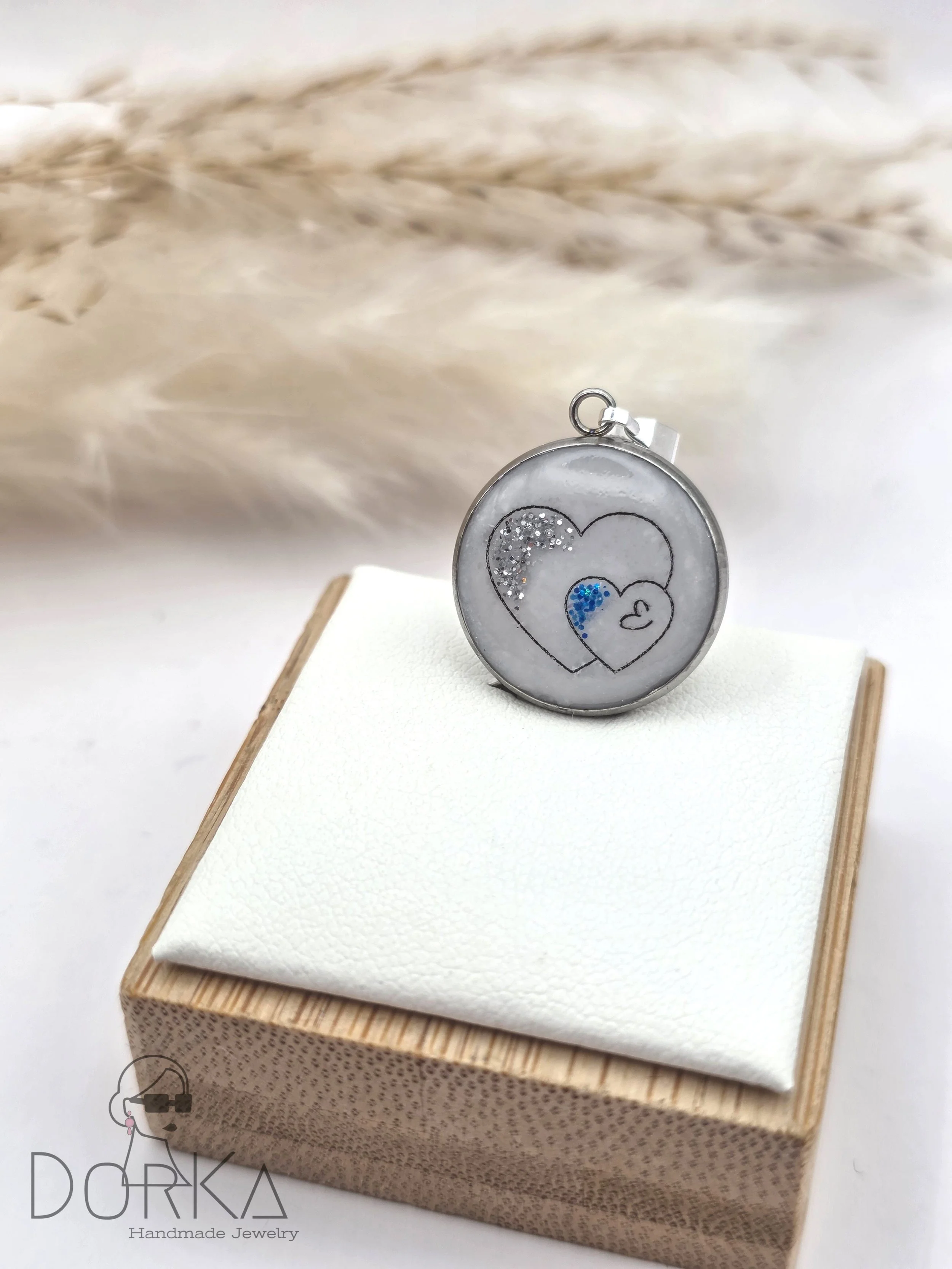 Ein runder Anhänger mit einem Herzdesign und glitzernden Elementen, auf einem weißen Stoff auf einem Holzständer präsentiert.