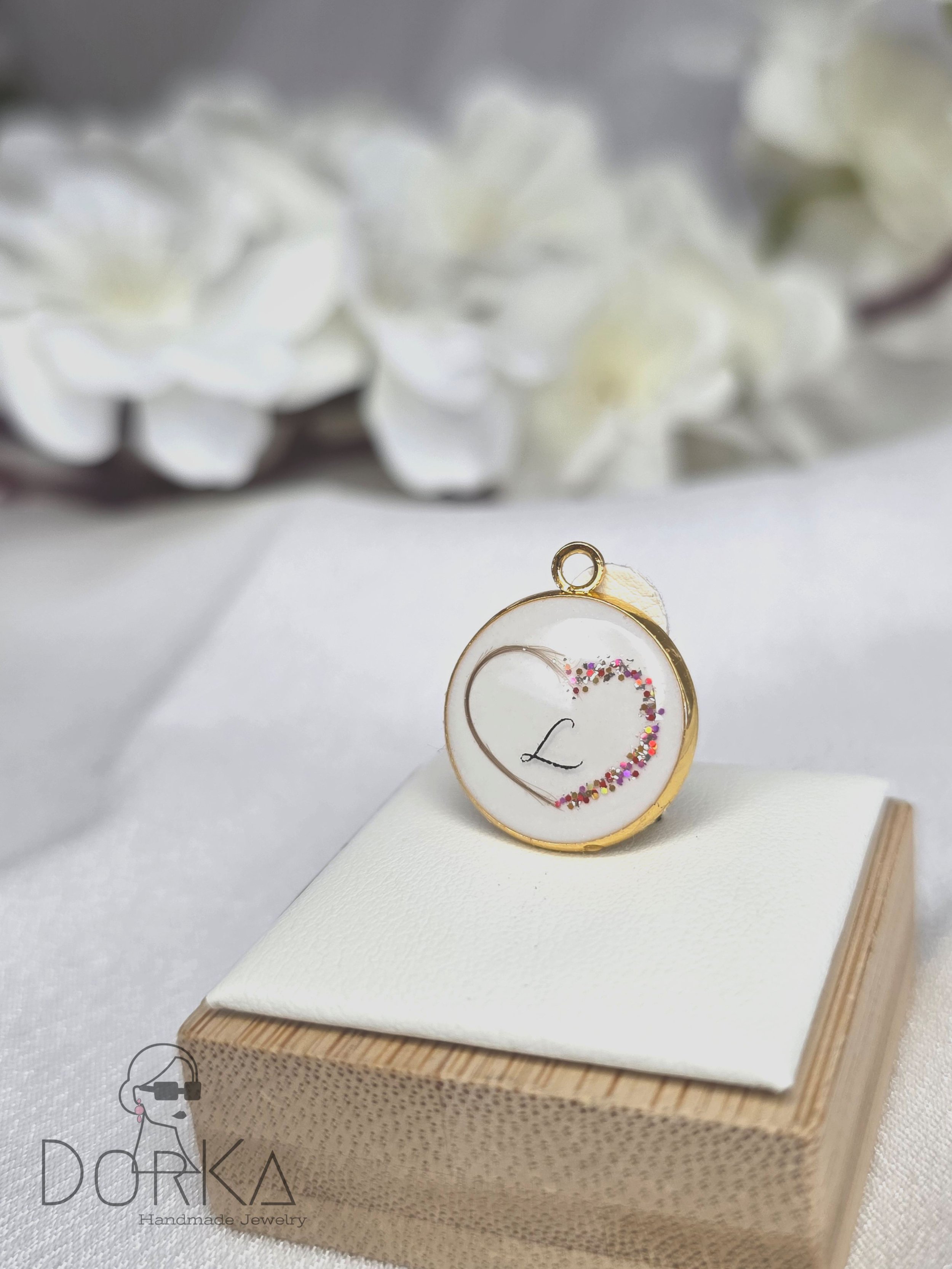 Ein runder Goldanhänger mit einer weißen Einlage, auf der ein Herz und der Buchstabe 'L' zu sehen sind, verziert mit buntem Glitter, auf einem weißen Stoffkissen auf einem Holzsockel, im Hintergrund weiße Blumen.