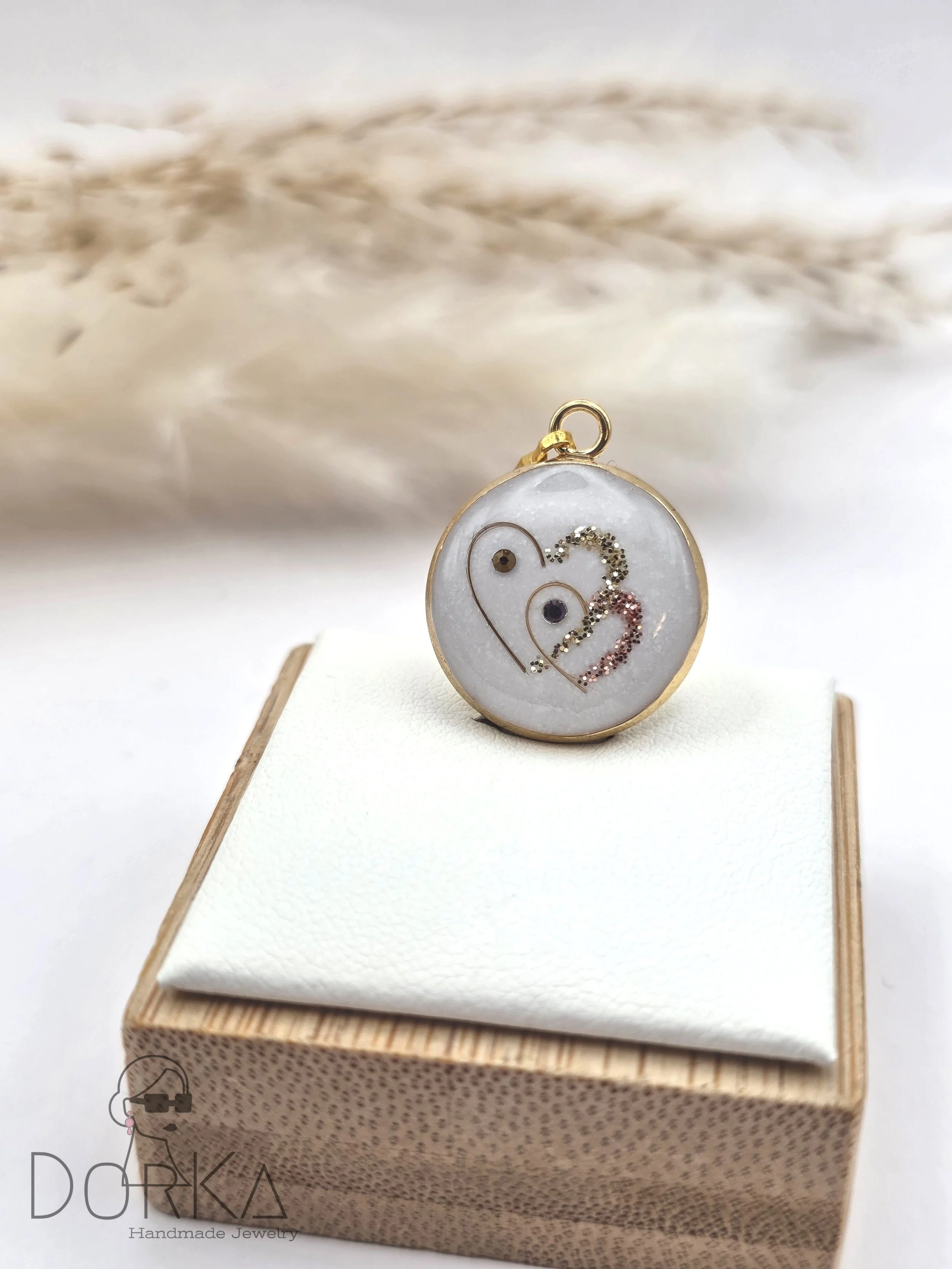 Ein runder, verschlüsselter Anhänger mit goldener Umrahmung, der auf einem weißen Kissen auf einer Holzkiste liegt. Der Anhänger enthält dekorative Elemente mit zwei kleinen schwarzen Steinen, die wie Augen wirken, und stilisierten Herzen aus kleinen