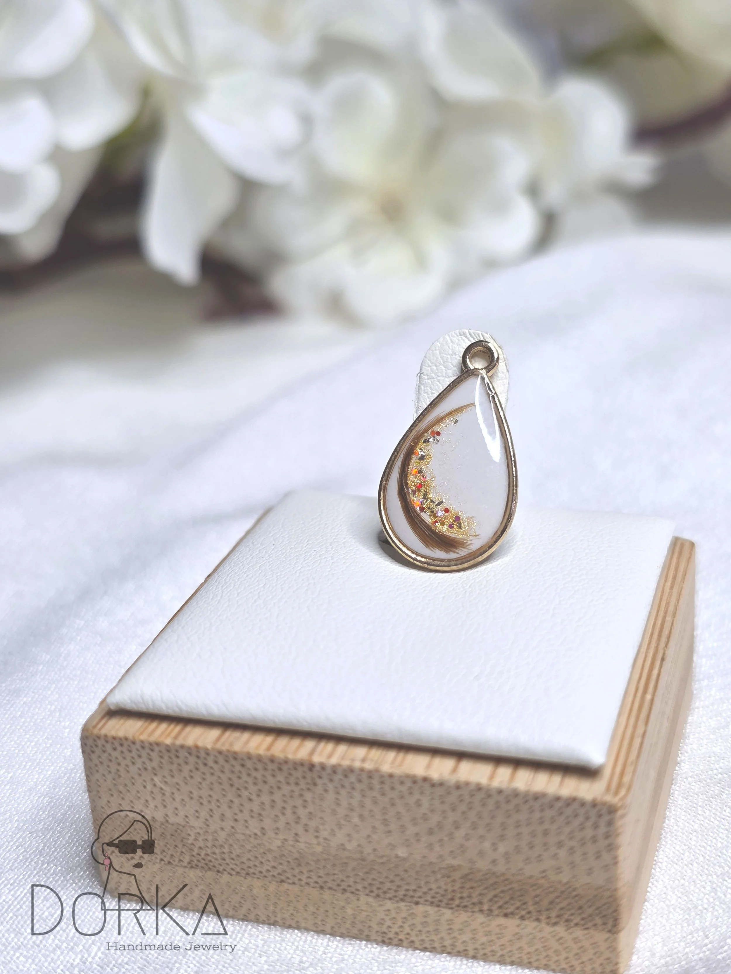 Ein Ohrring mit Wassertropfen-Design, verziert mit bunten Glitzer-Partikeln, auf einem weißen Kissen, auf einem Holzsockel, im Hintergrund weiße Blumen.