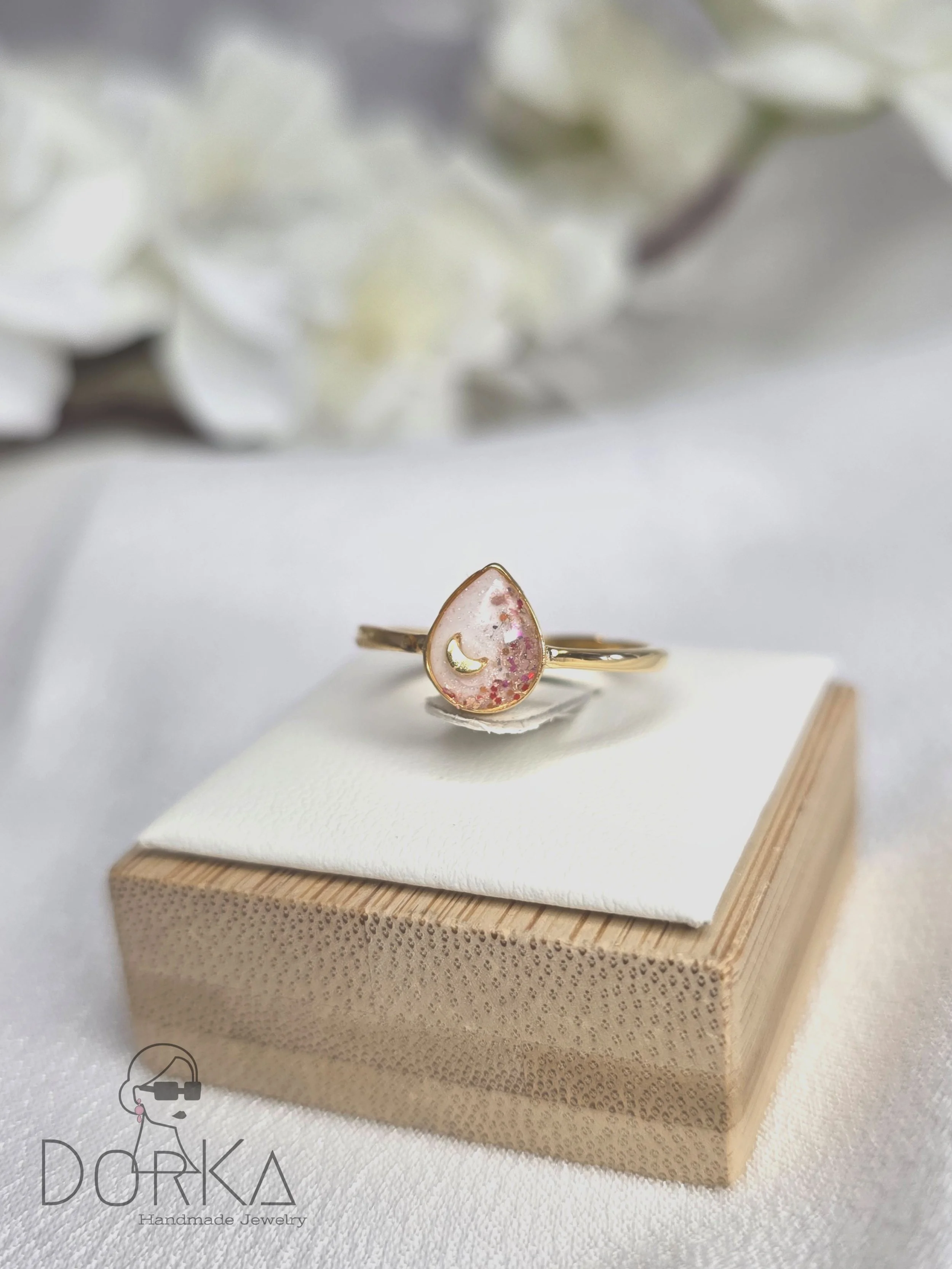 Ein Goldring mit einem pinken, tröpfchenförmigen Anhänger, der kleine Glitzerpartikel und eine goldene Mondsichel enthält, liegt auf einer kleinen Holzplatte.