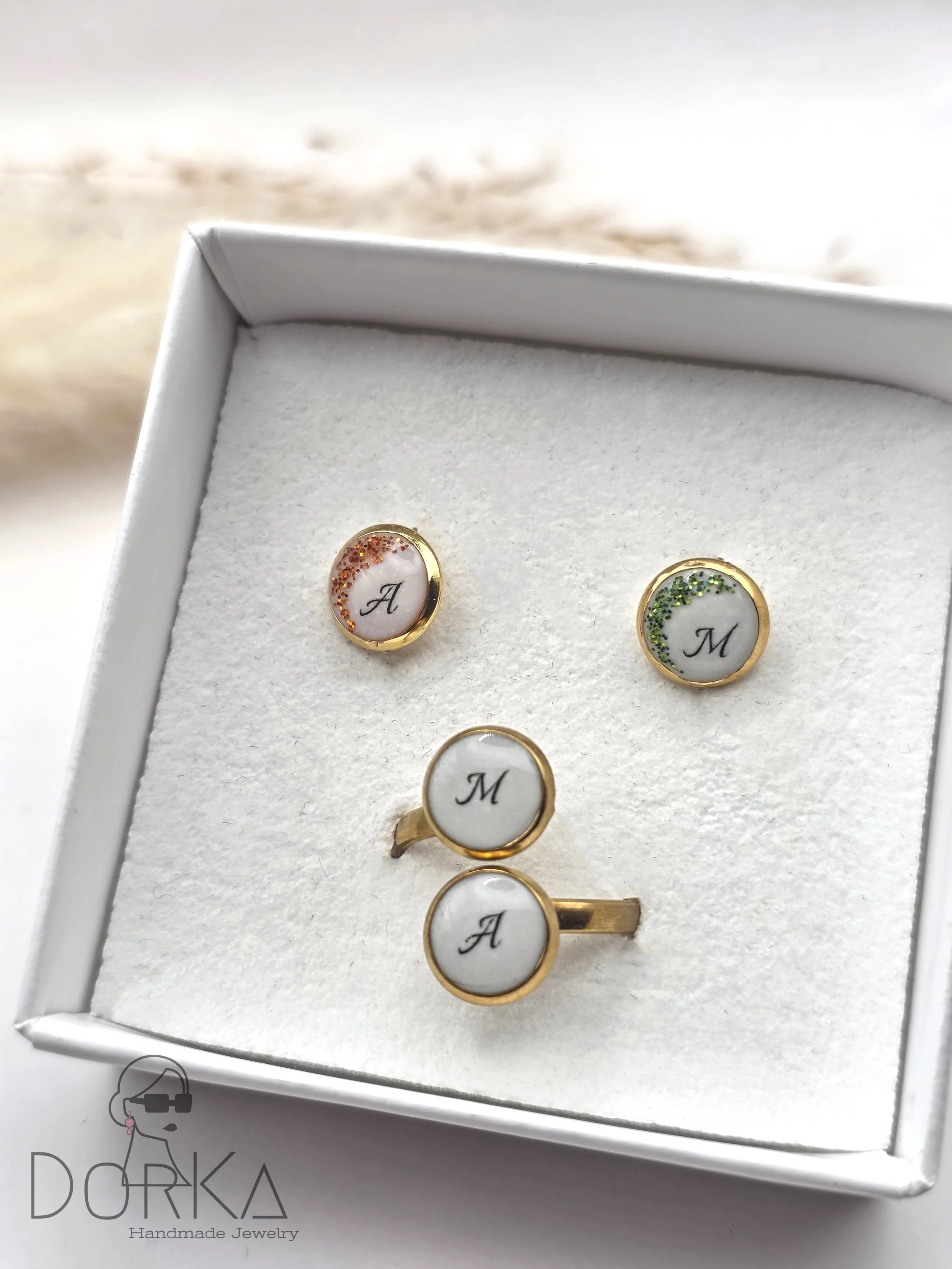 Handgemachte Schmuckset, bestehend aus einem Ring und zwei Ohrsteckern mit weißen ovalen Steinen, darauf schwarz gedruckte Buchstaben ‚A‘ und ‚M‘, in einer weißen Geschenkbox mit Logo ‚DORKA Handmade Jewelry‘