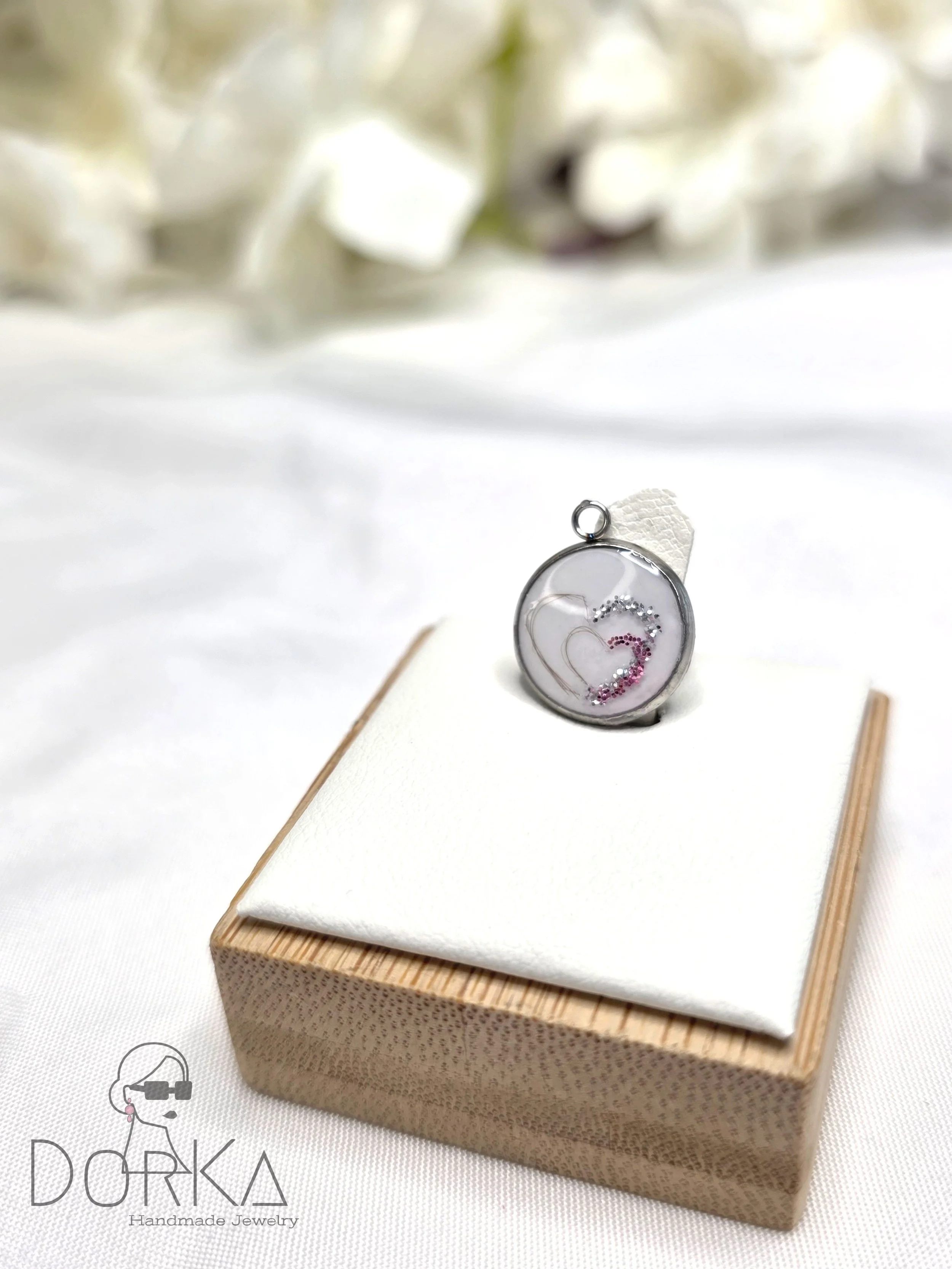 Eleganter handgemachter Anhänger mit Herzdesign auf einer weißen Schachtel, im Hintergrund weiße Blumen.