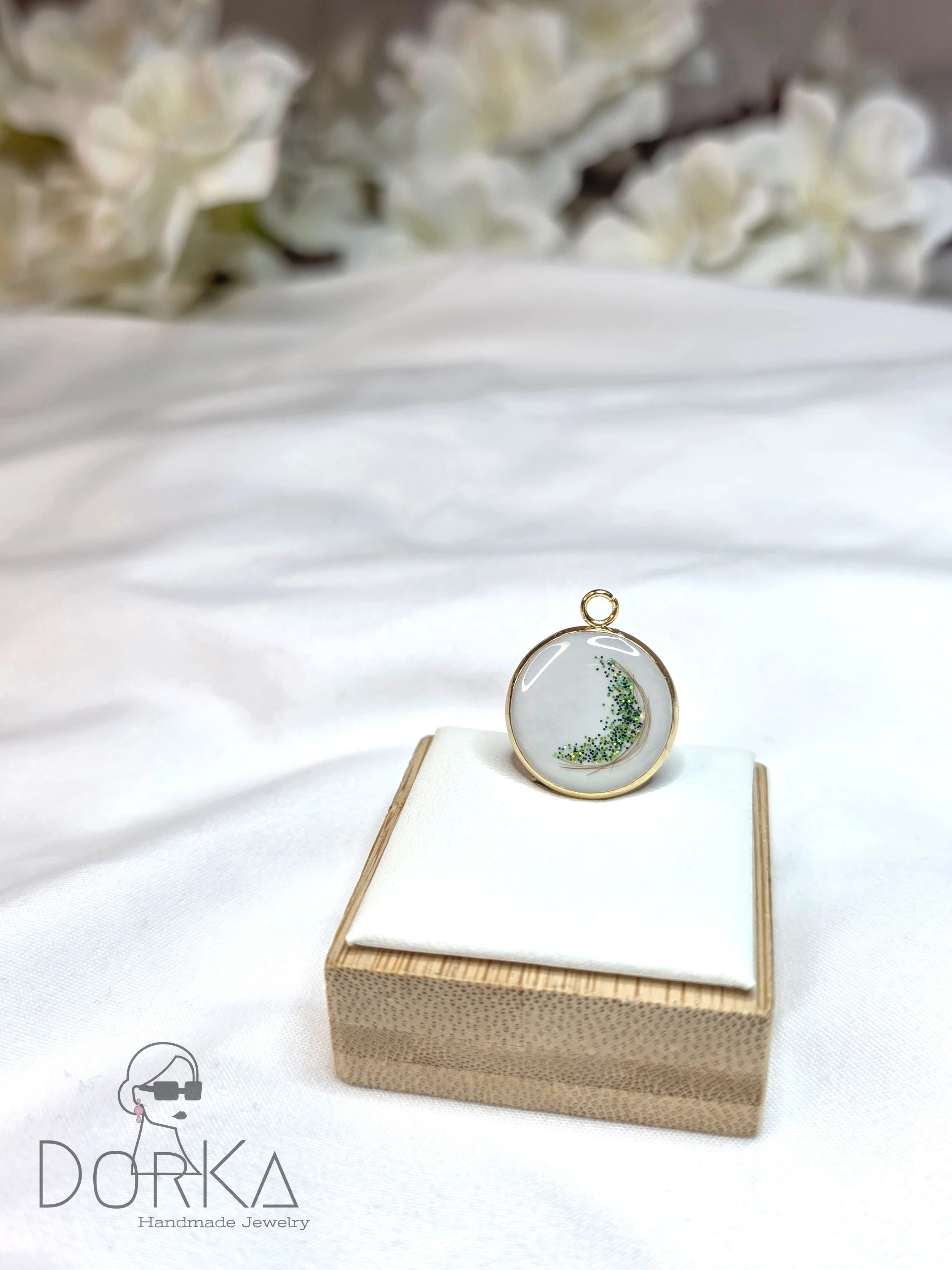 Ein runder Anhänger mit einem kleinen Mond aus glitzerndem grünen Glitter, in einem goldfarbenen Rahmen, auf einem weißen Kissen auf einem Holzetui. Im Hintergrund weiße Blumen.