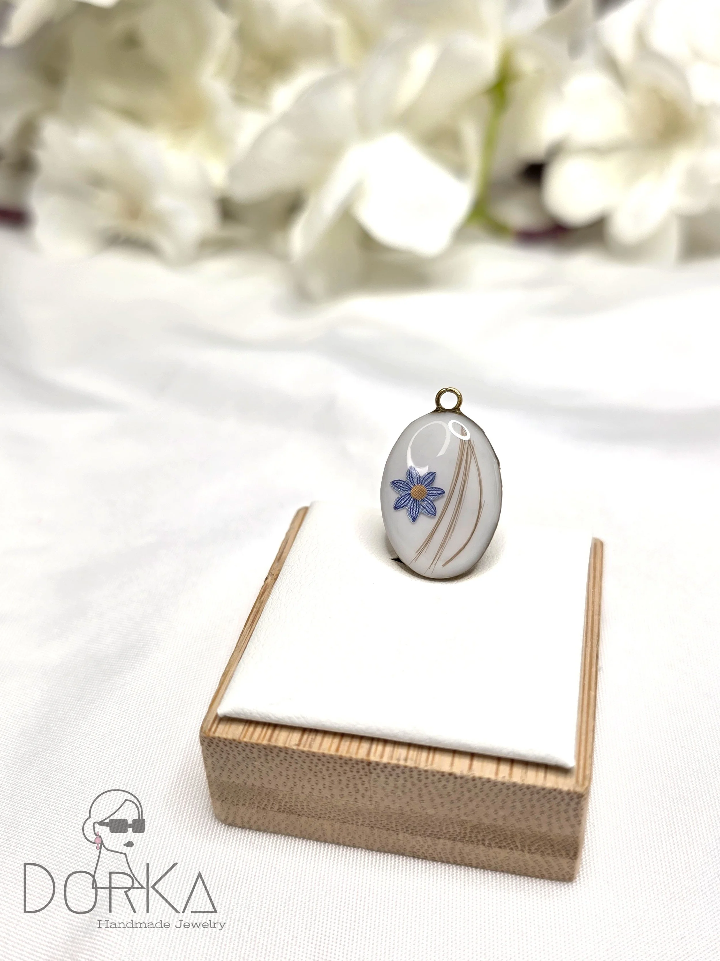 Ein handgemachtes Schmuckstück, ein ovaler Anhänger mit blauer Blume und goldenen Linien, auf einem weißen Stofftuch auf einem kleinen Holzsockel, im Hintergrund weiße Blumen.