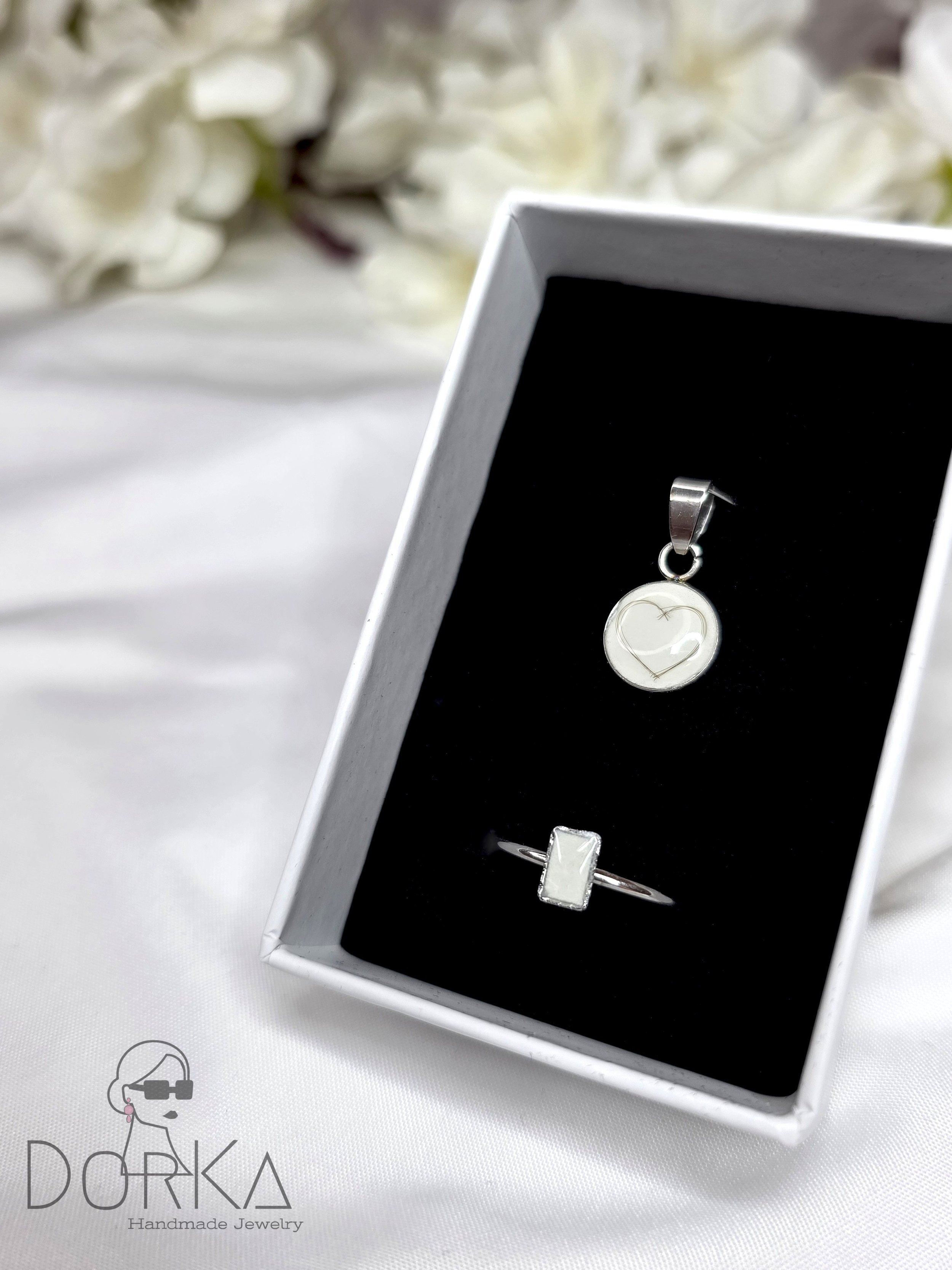 Silberne Schmuckstücke in einer schwarzen Geschenkbox, bestehend aus einem Anhänger mit Herz-Design und einem Ring mit rechteckigem Edelstein, auf weißem Hintergrund mit weißen Blumen im Hintergrund.