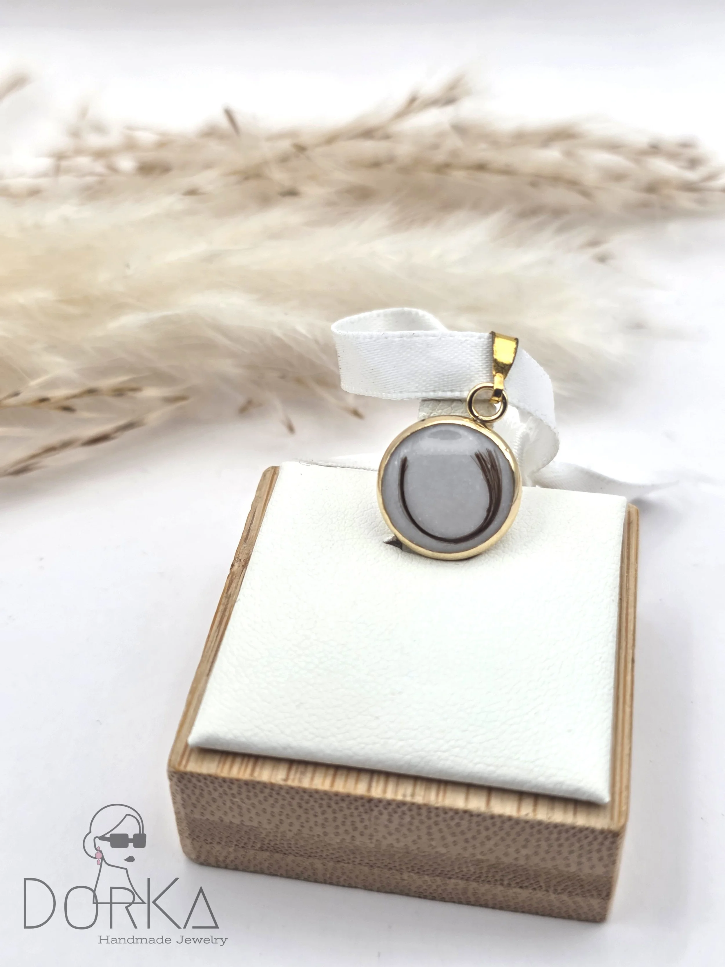 Ein handgefertigtes Schmuckstück, ein Anhänger mit einem grafischen Kreisdesign, liegt auf einem weißen Kissen in einer runden Holzschatulle. Im Hintergrund sind getrocknete Gräser.