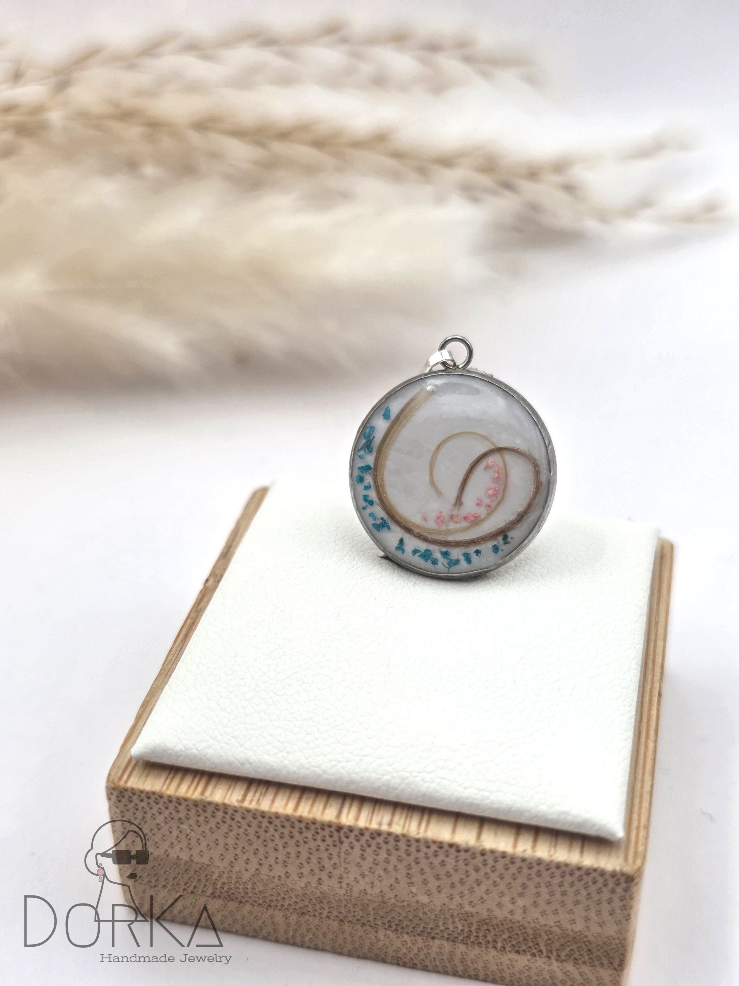 Rundes, handgemachtes Schmuckstück mit spiralförmigem Design, in einem transparenten Rahmen auf einem weißen Kissen, auf einem kleinen Holzständer präsentiert.