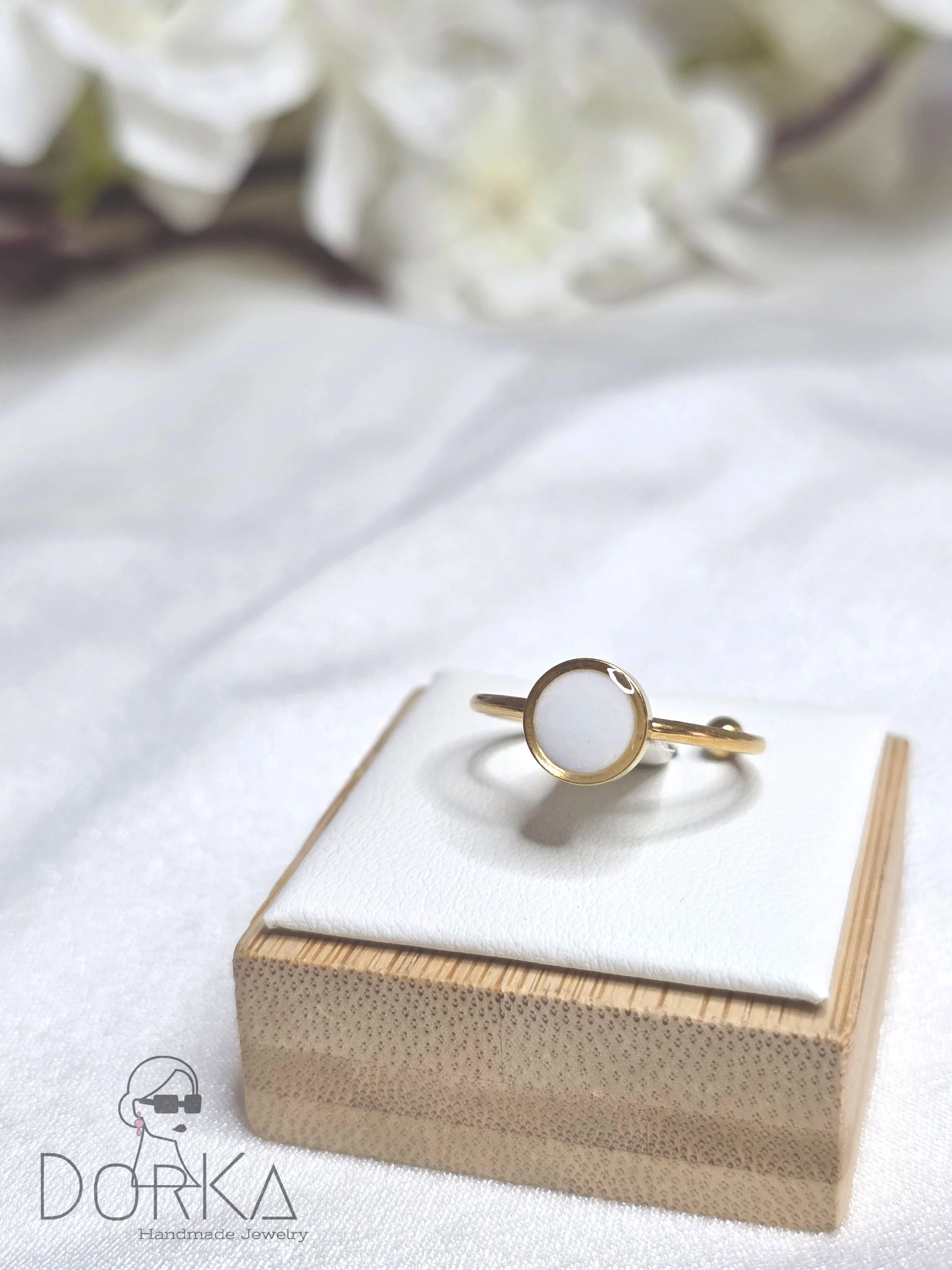 Goldener Ring mit weißem Stein auf weichem weißen Stoff, im Hintergrund weiße Blumen.