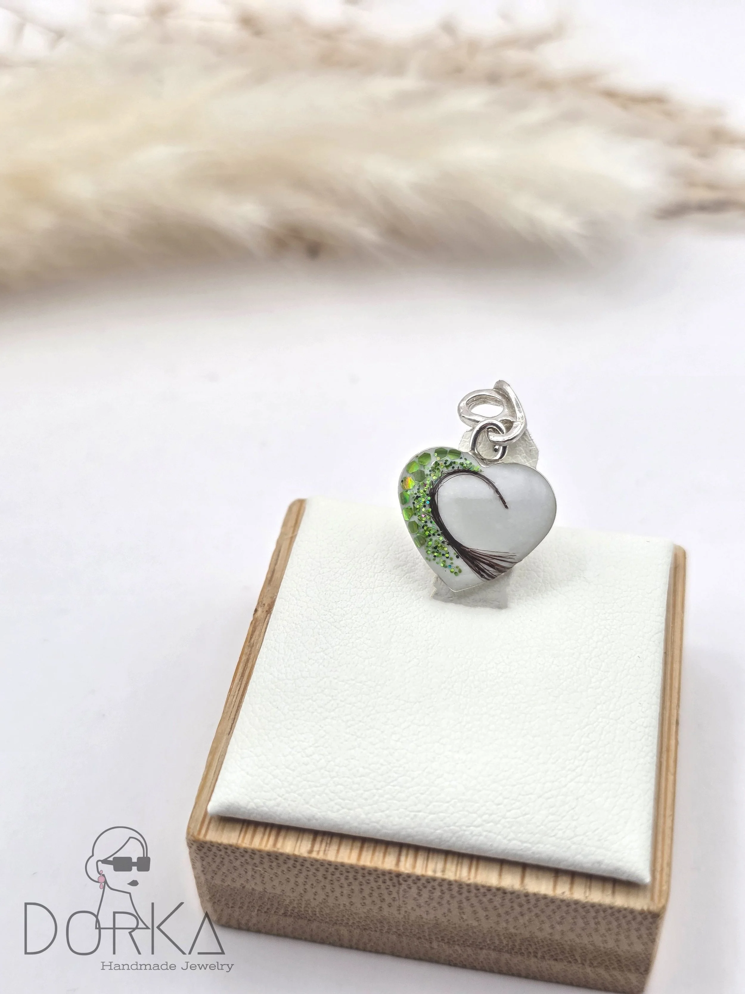 Ein silbernes Herz-Anhänger mit weißen und grünen Akzenten, ausgeführt mit einem braunen Linienmuster, auf einem weißen Schmuckkissen in einer kleinen Holzschachtel, mit dem Logo „Dorka Handmade Jewelry“ im unteren linken Eck.