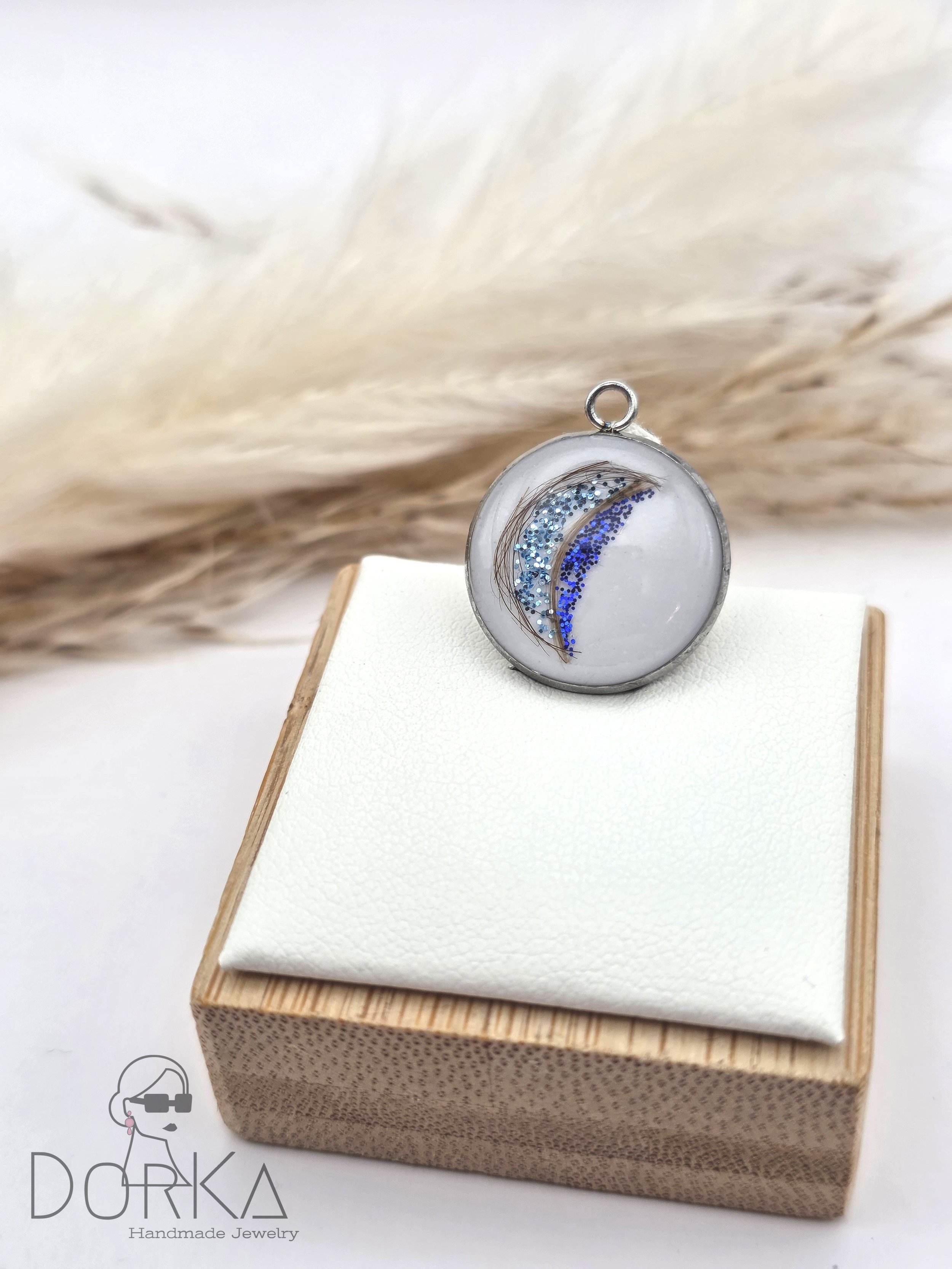 Ein handgefertigtes Schmuckstück, ein runder Anhänger mit blauer Glitzer und Federn, auf einem weißen Kissen auf einer Holzkiste.