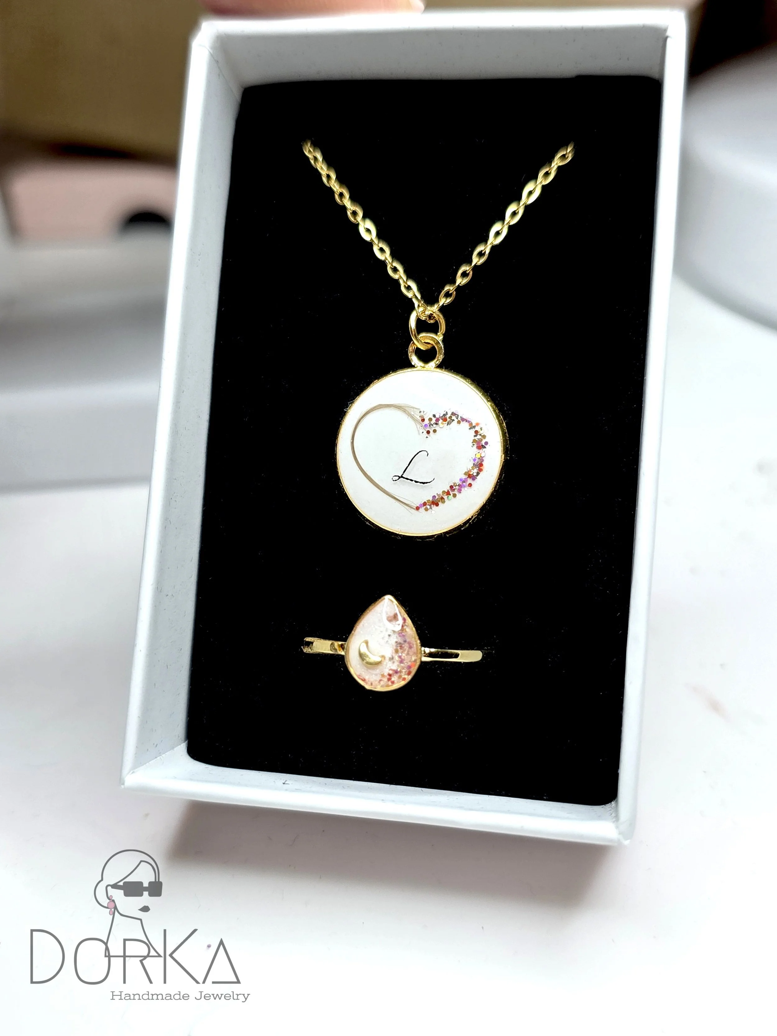 Goldene Halskette mit Anhänger, der ein Herz mit Schleife und bunten Glitzer enthält, sowie einen goldenen Ring mit tropfenförmigem Anhänger, der pinken und goldenen Glitzer aufweist, in einer schwarzen Schmuckschachtel.