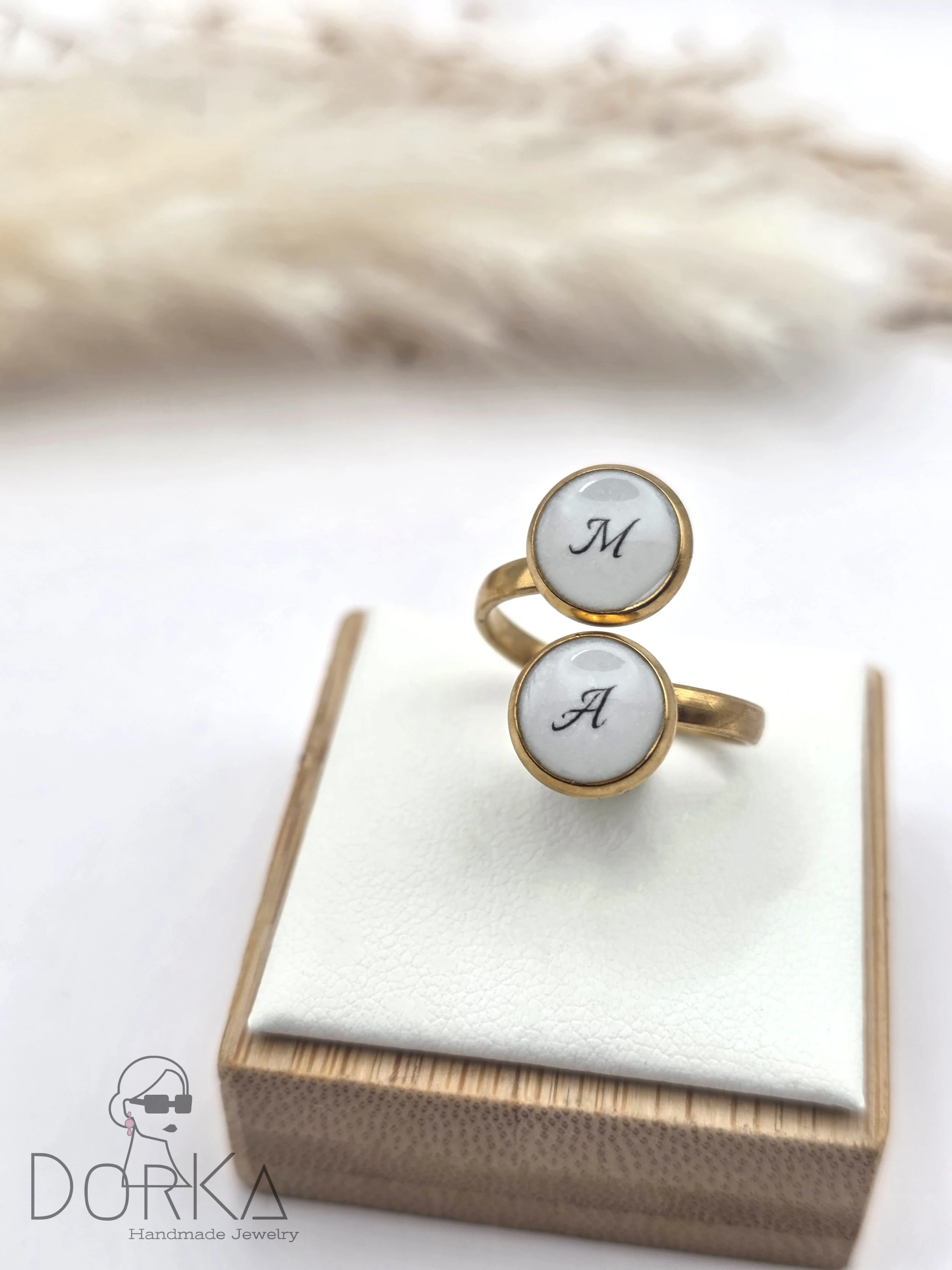 Goldenes Ring mit weißen Zifferblättern, auf denen die schwarzen Buchstaben 'M' und 'A' stehen, auf weißer Samtunterlage in einer kleinen Schmuckschatulle.