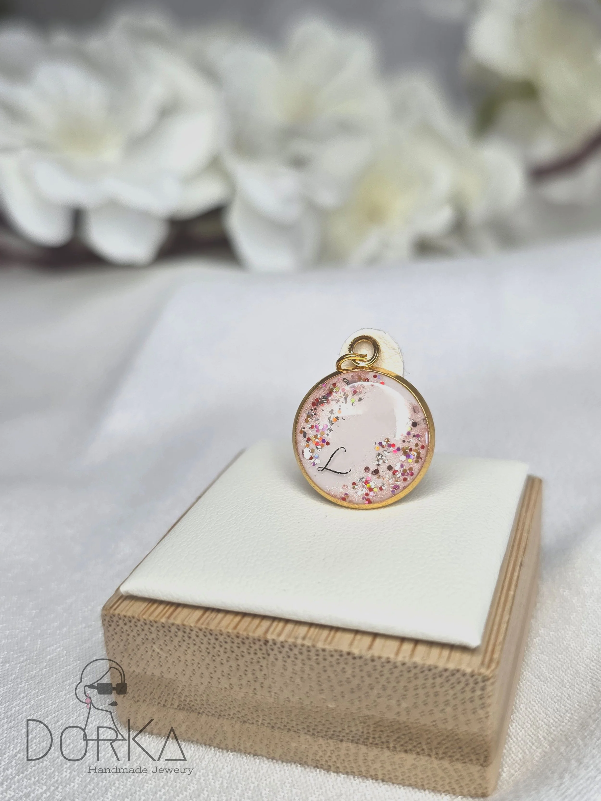Ein runder Anhänger mit rosa Glitzer und einem eingedruckten Buchstaben 'L', aufgestellt auf einem weißen Kissen auf einem Holzkästchen, im Hintergrund weiße Blumen.