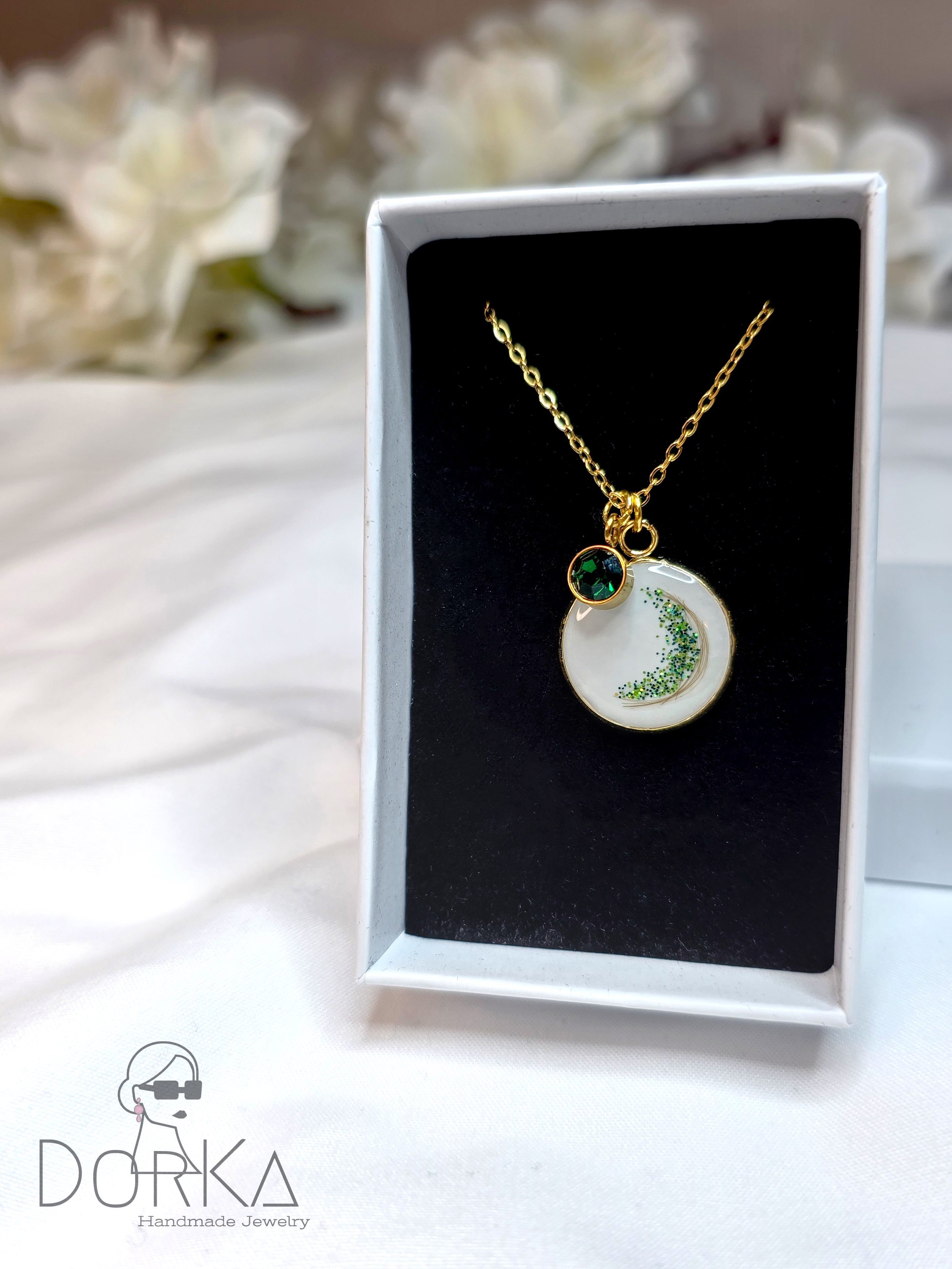 Goldene Halskette mit Anhänger in Form eines Halbmonds mit grünen Glitzerpartikeln und einem grünen Stein, präsentiert in einer schwarzen Schmuckhalterung in einer weißen Schmuckschachtel, im Hintergrund weiße Blumen.
