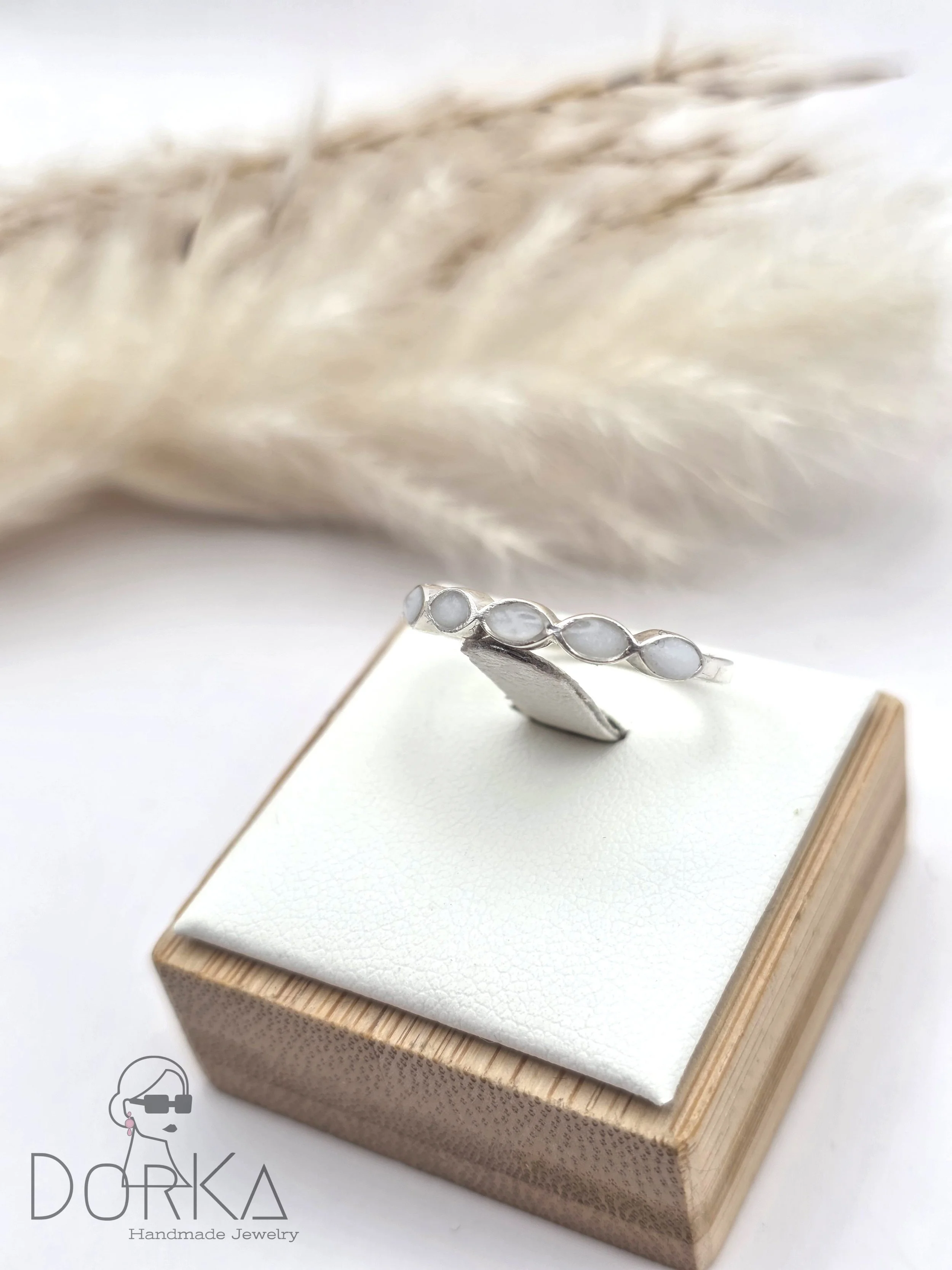 Silberner Ring mit ovalen weißen Steinen auf einem weißen und Holzschatullen. Im Hintergrund sind weiche, helle Pflanzen. Das Bild trägt das Logo 'DORKA Handmade Jewelry'.
