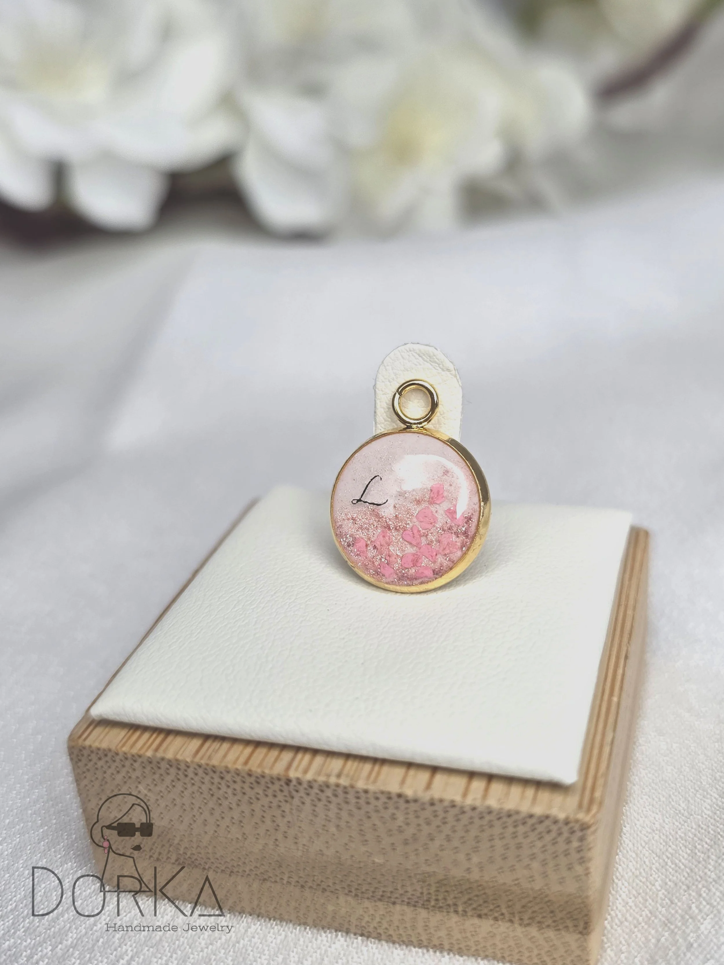 Ein goldener Ohrring mit einem transparenten Anhänger, in dem rosa Blütenblätter und der Buchstabe 'L' enthalten sind, präsentiert auf einem weißen Kissen in einer Holzschachtel, im Hintergrund weiße Blumen.