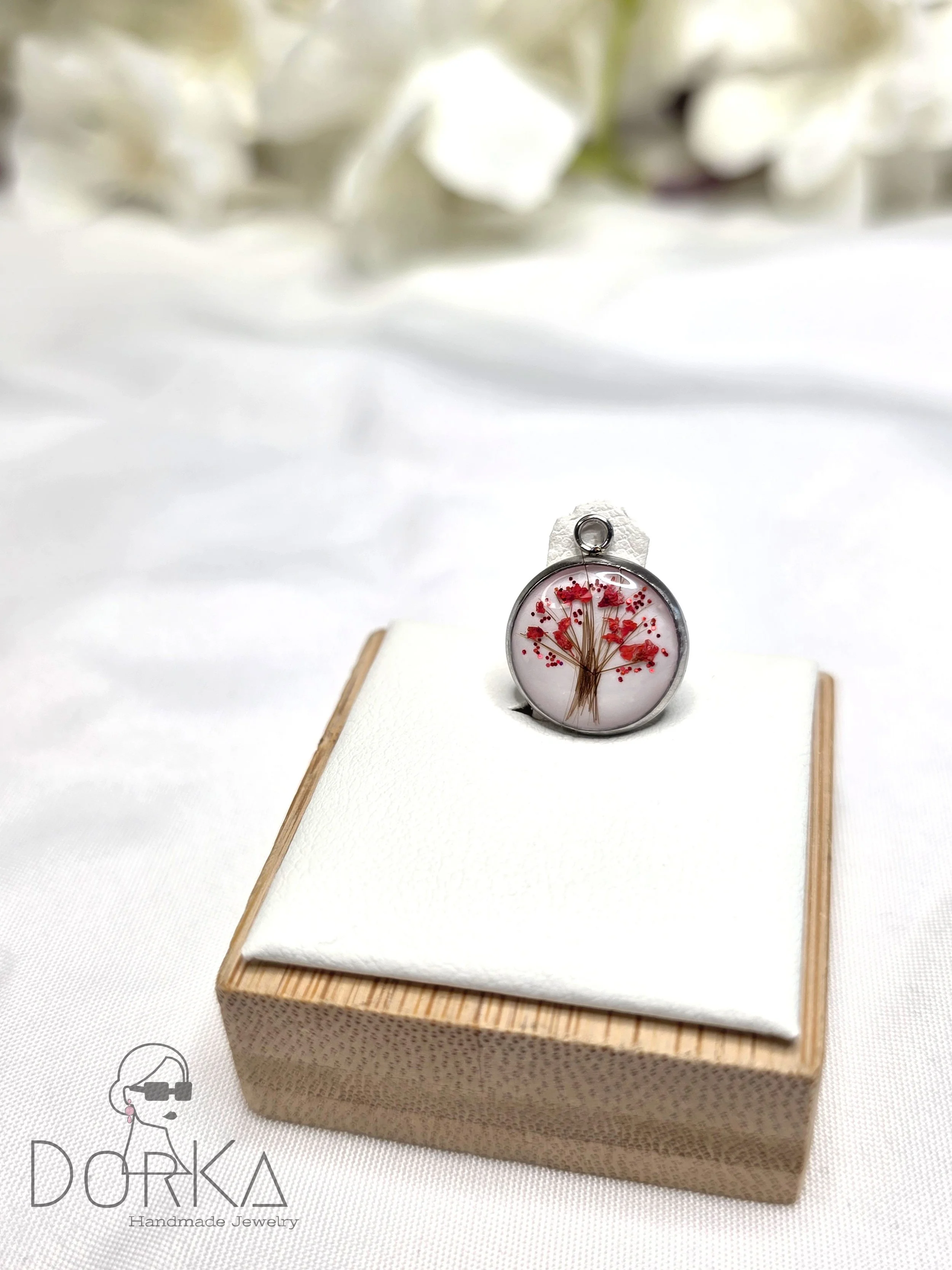 Ein runder Anhänger mit einem eingearbeiteten, getrockneten Blumenmuster in roten und braunen Tönen, auf einem weißen Stoff auf einem kleinen Holzkästchen präsentiert.