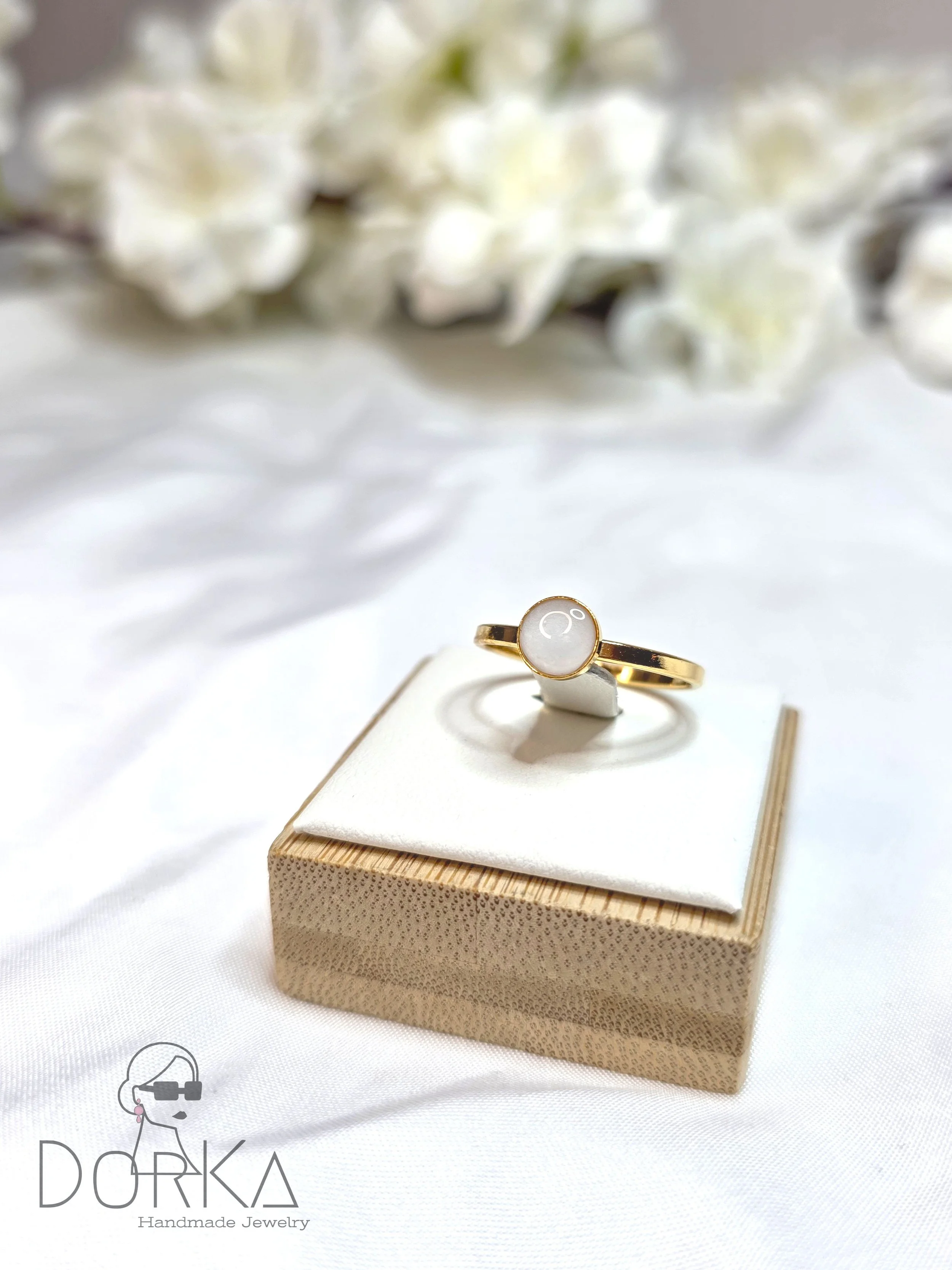 Ein goldener Ring mit einem weißen, runden Edelstein auf einem weißen Sockel, vor einem weißen Blumenkleid. Das Logo "DORKA Handmade Jewelry" ist im unteren linken Eck.