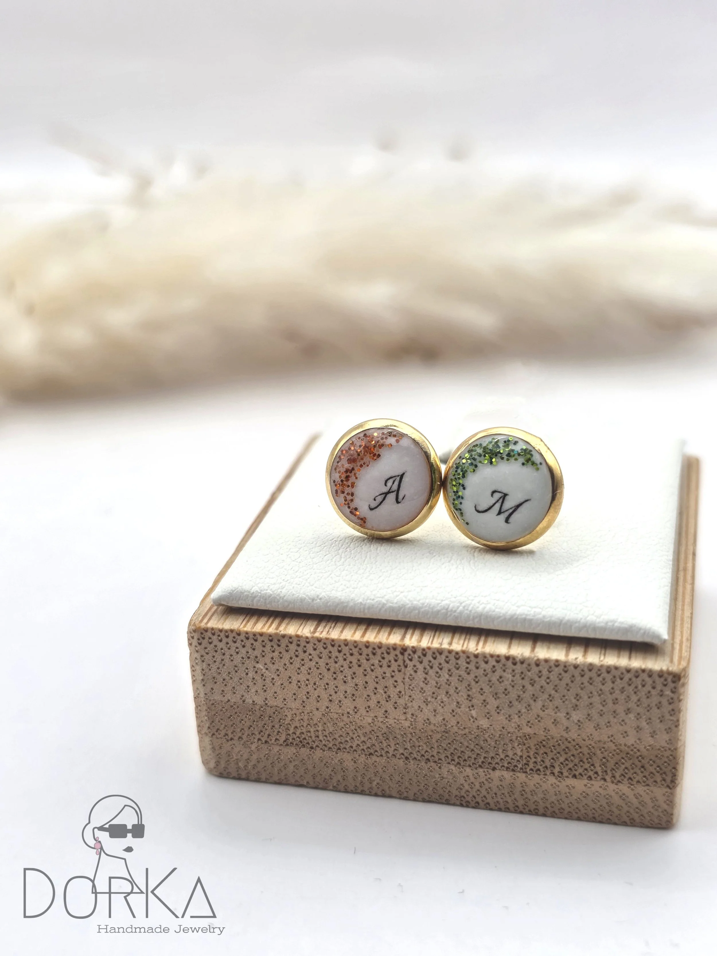 Ein Paar handmade, personalisierte Manschettenknöpfe mit den Buchstaben 'A' und 'M', verziert mit bunten Glitter, auf einem weißen Kissen, das auf einem Holzkästchen liegt.