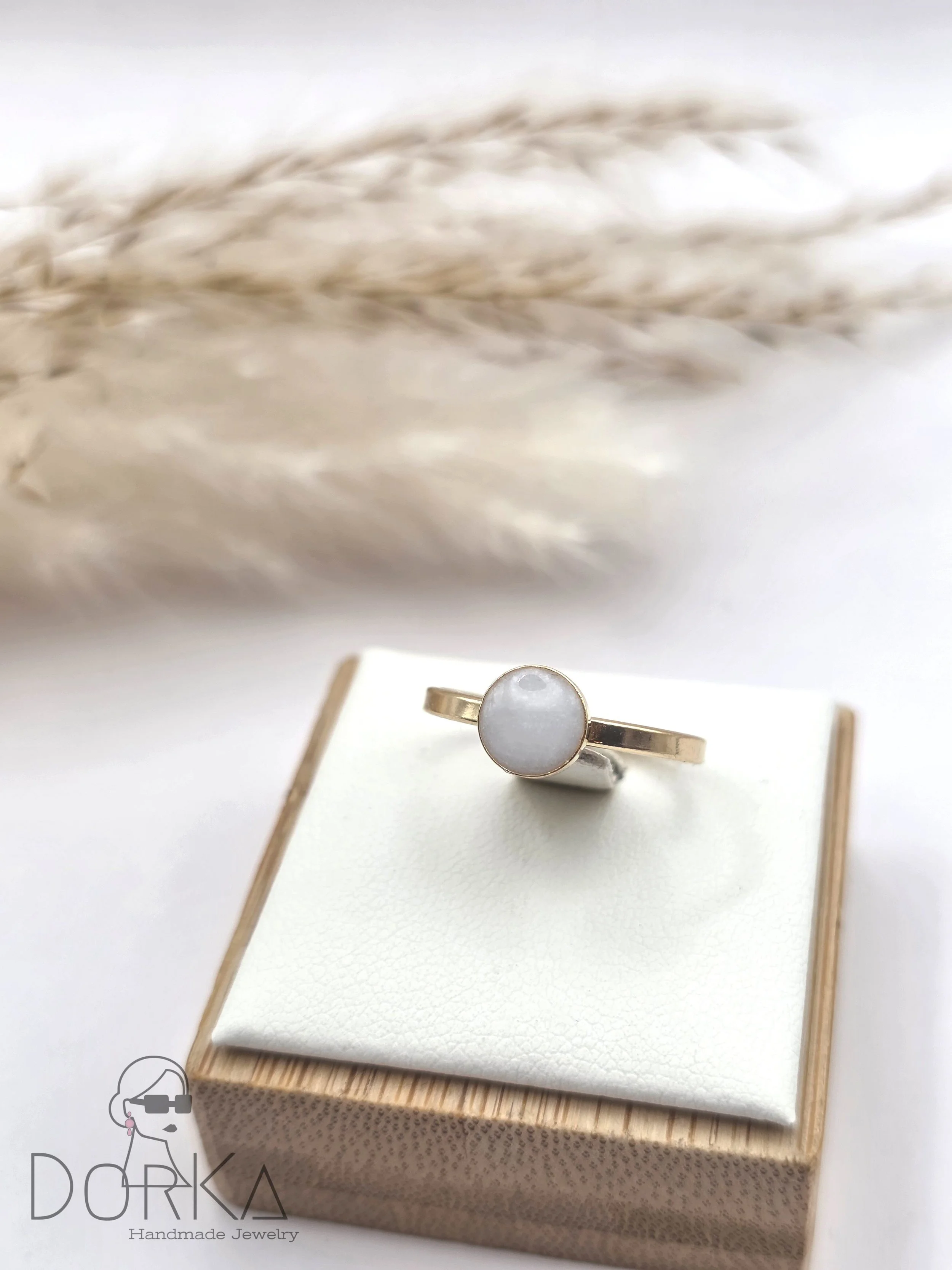 Goldring mit weißem ovalem Edelstein auf silberner Ständer auf Geschenkbox, im Hintergrund getrocknete Gräser