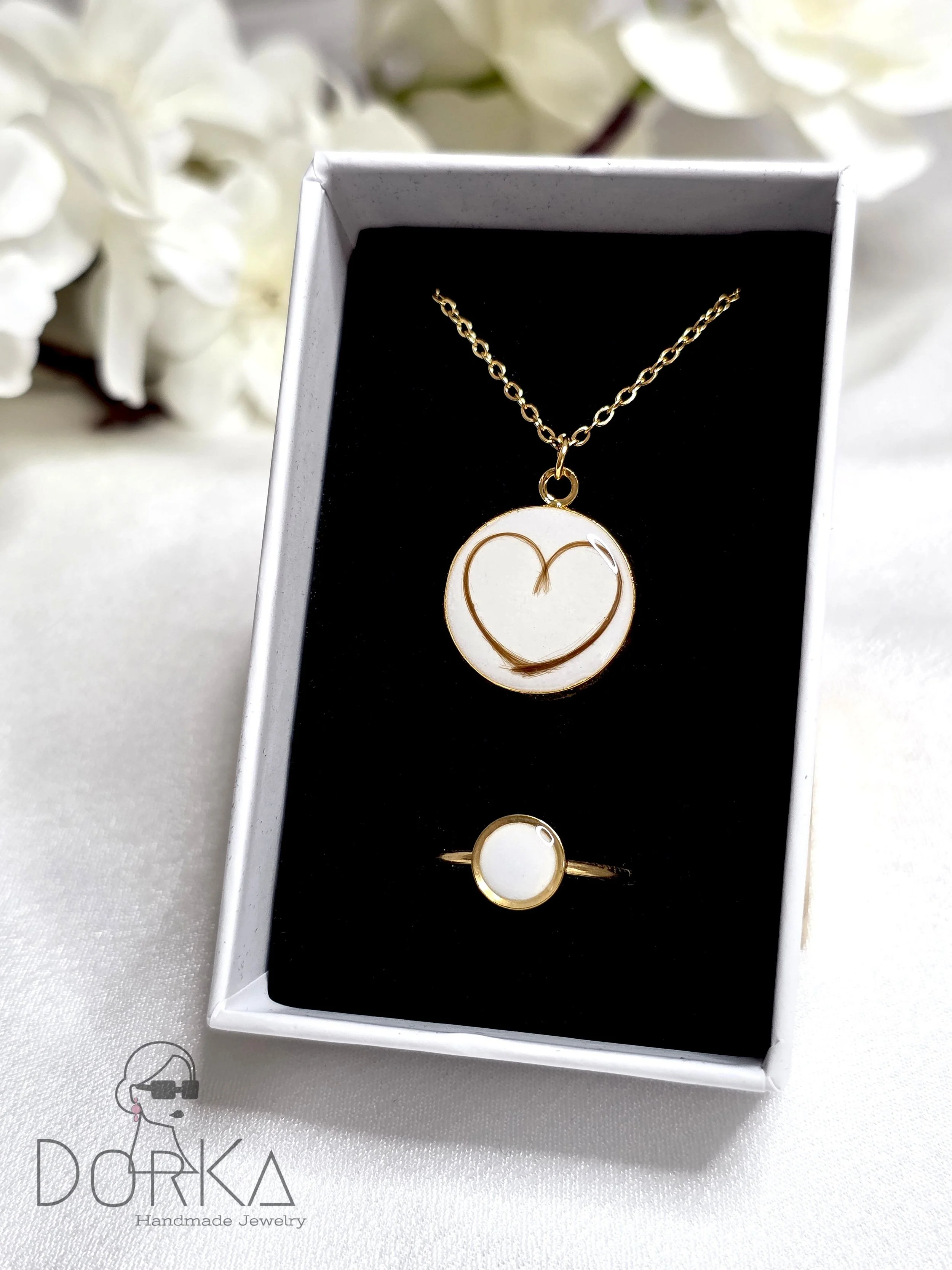 Ein Schmuckset bestehend aus einer goldenen Halskette mit einem weißen Anhänger in Herzform und einem goldenen Ring mit weißem Stein, in einer schwarzen Schmuckbox. Im Hintergrund weiße Blumen.