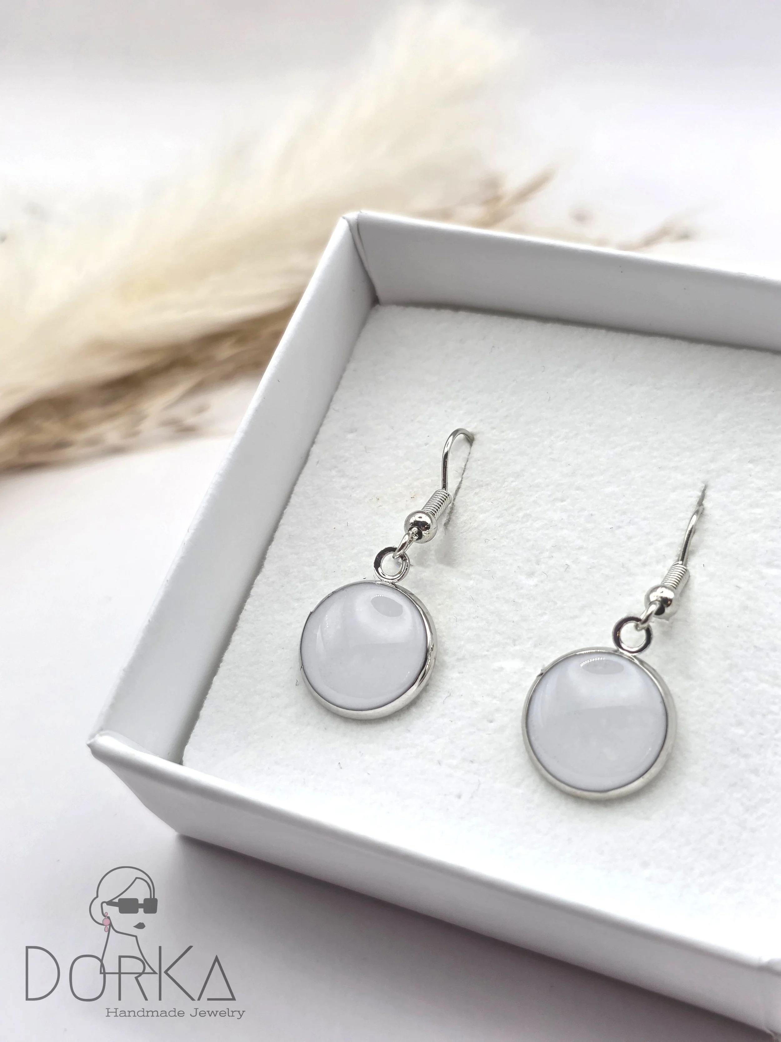 Ein Paar silberne Ohrhänger mit weißen, runden Steinen in einer weißen Schmuckschachtel, Dorka Handmade Jewelry, im Hintergrund ein heller, kuscheliger Stoff.
