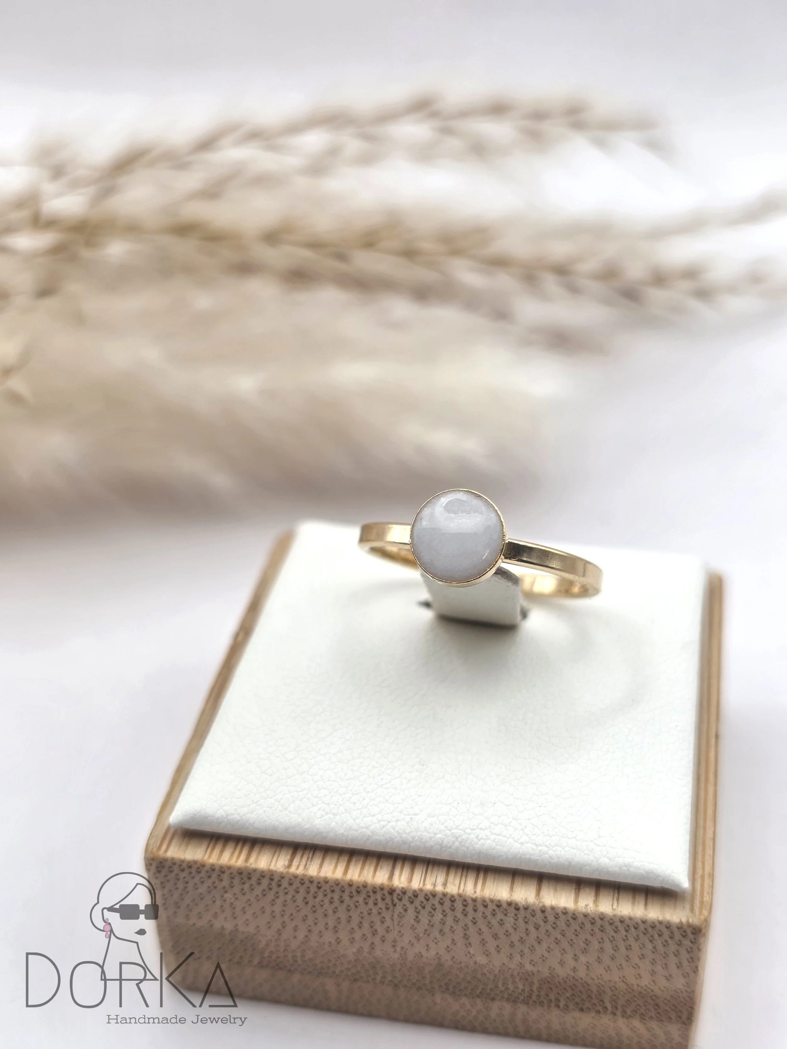 Goldring mit weißem, runden Edelstein auf einem Schmuckkissen. Im Hintergrund getrocknete Gräser, Logo "Dorka Handmade Jewelry" im unteren linken Bereich.