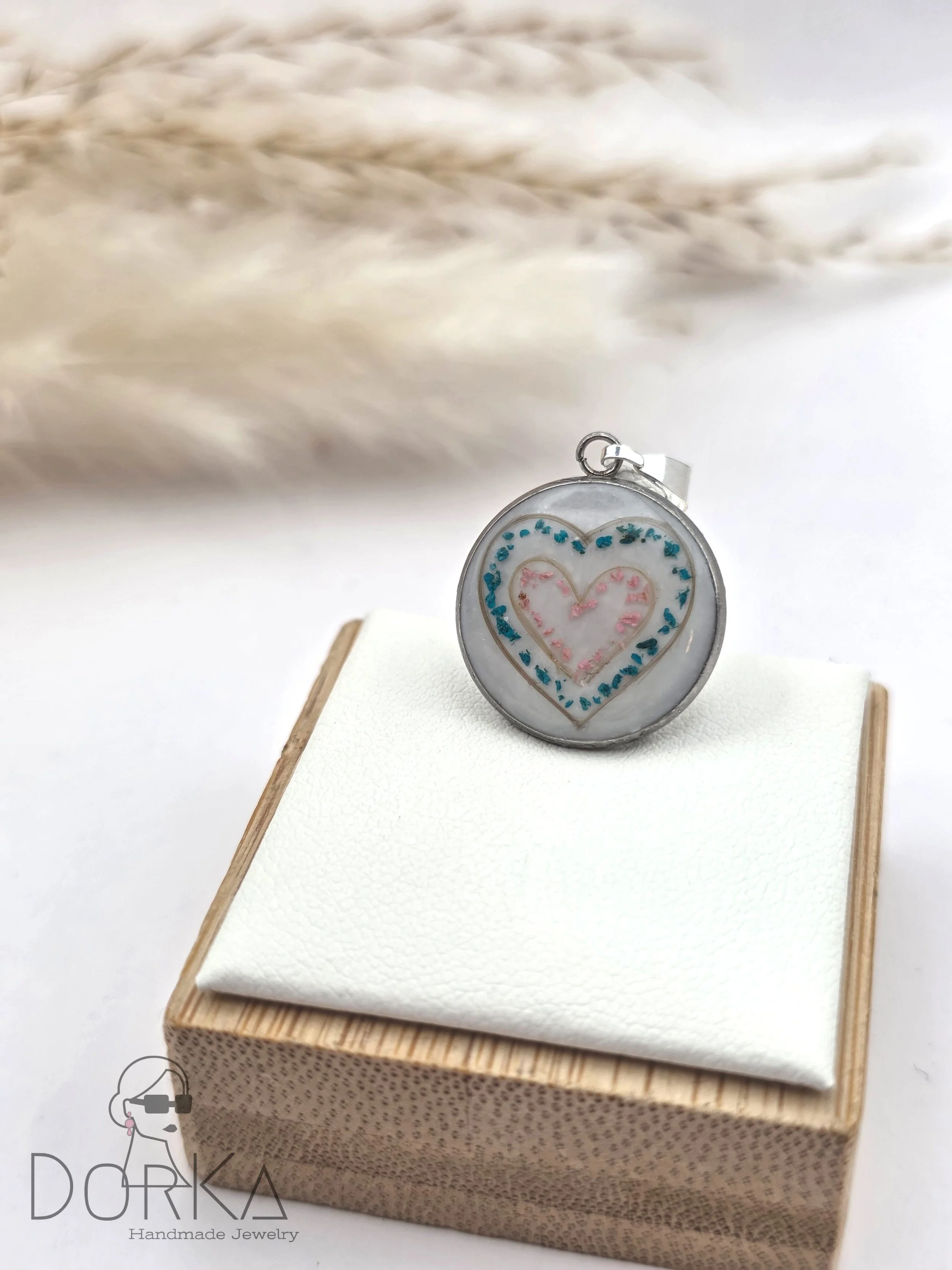 Ein runder Anhänger mit einem handgemalten, rosa und blauen Herz-Design, auf einem weißen Kissen auf einem kleinen Holzkästchen.