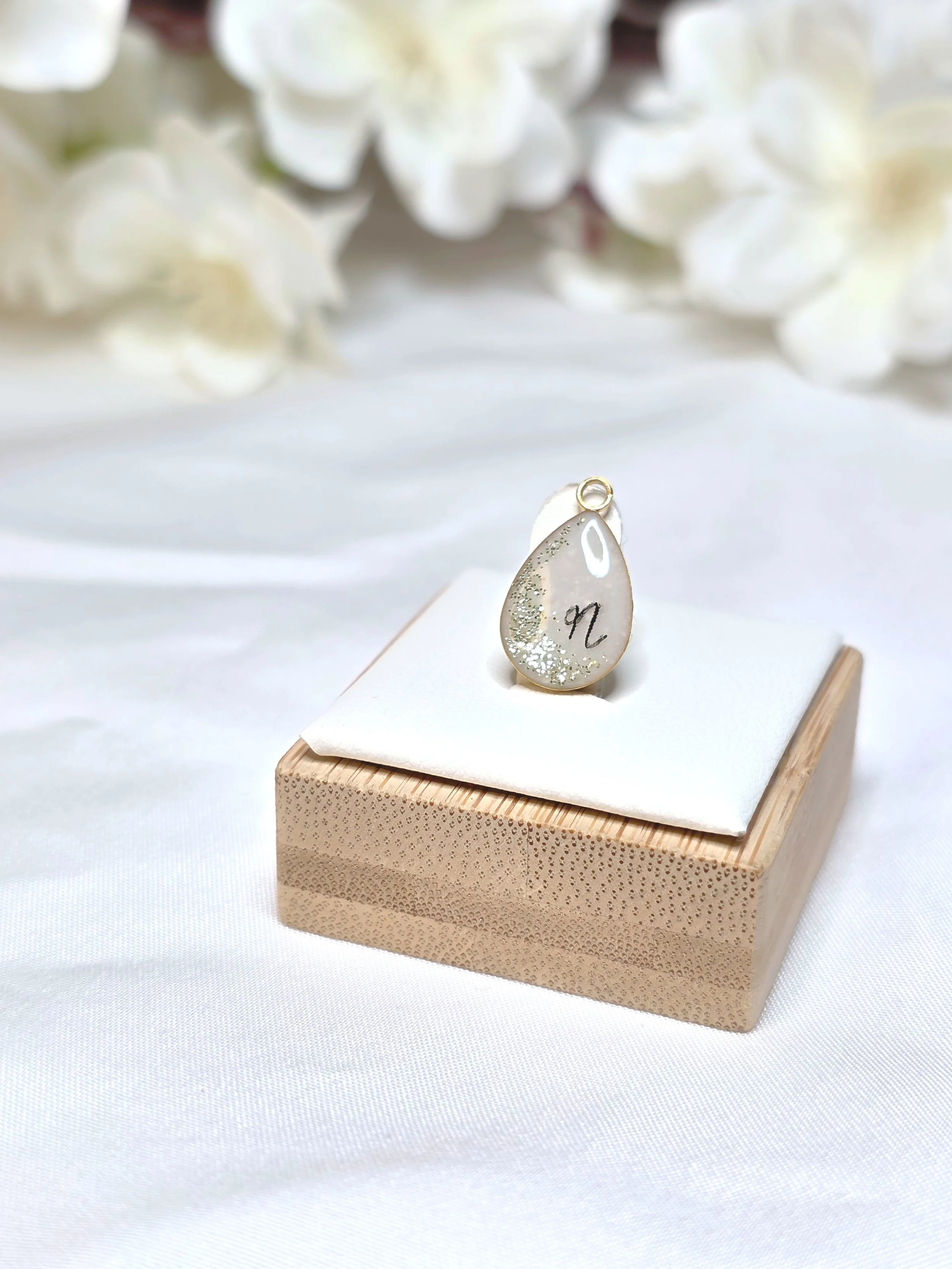 Ein Anhänger mit einer Perle und einem schwarzen Symbol auf einer weißen Unterlage auf einem Holztisch, im Hintergrund weiße Blumen.