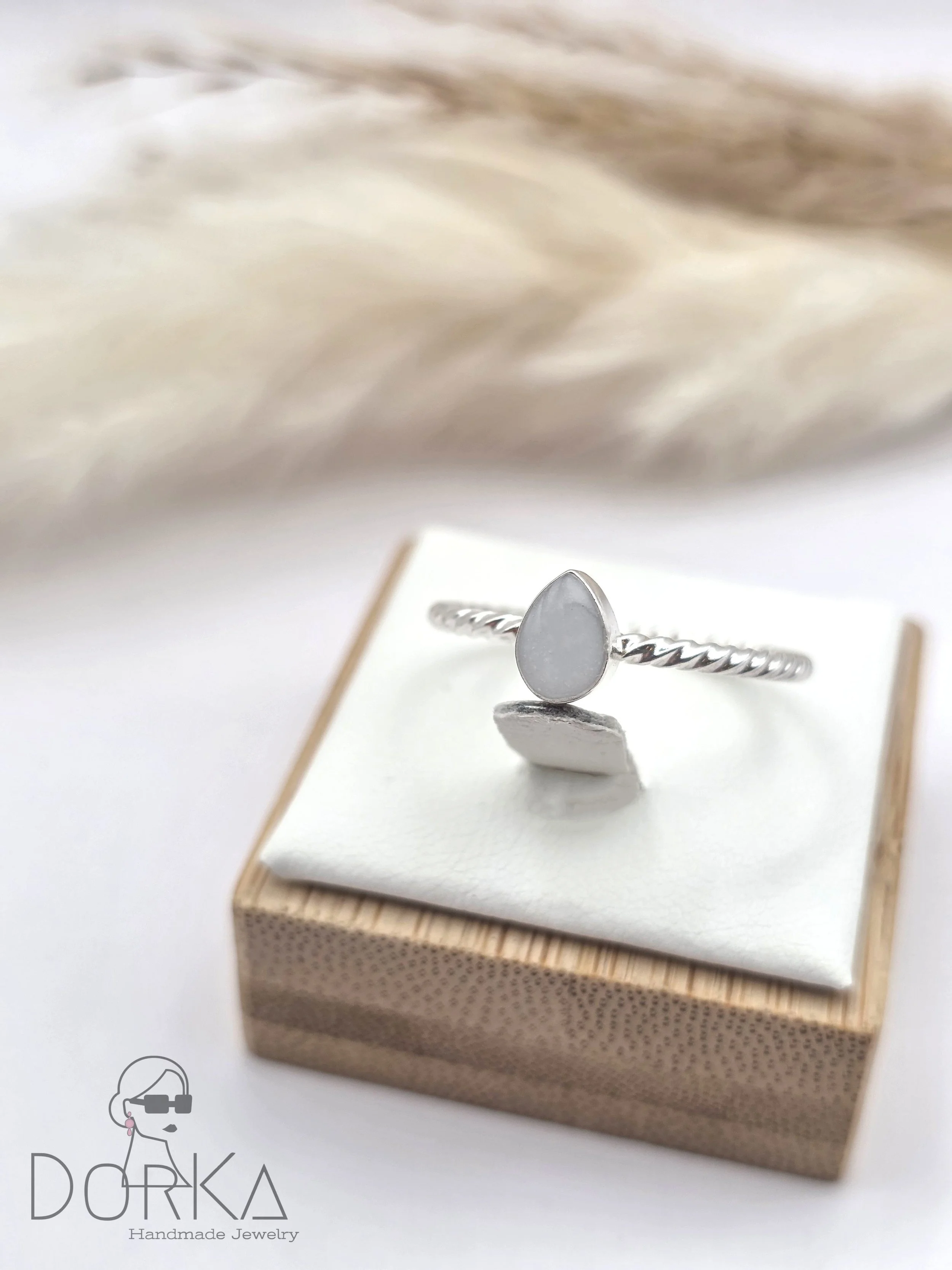 Silberring mit einem weißen, tropfenförmigen Edelstein auf einem weißen Kissen, auf einem kleinen Holzkästchen, im Hintergrund eine weiße Feder.