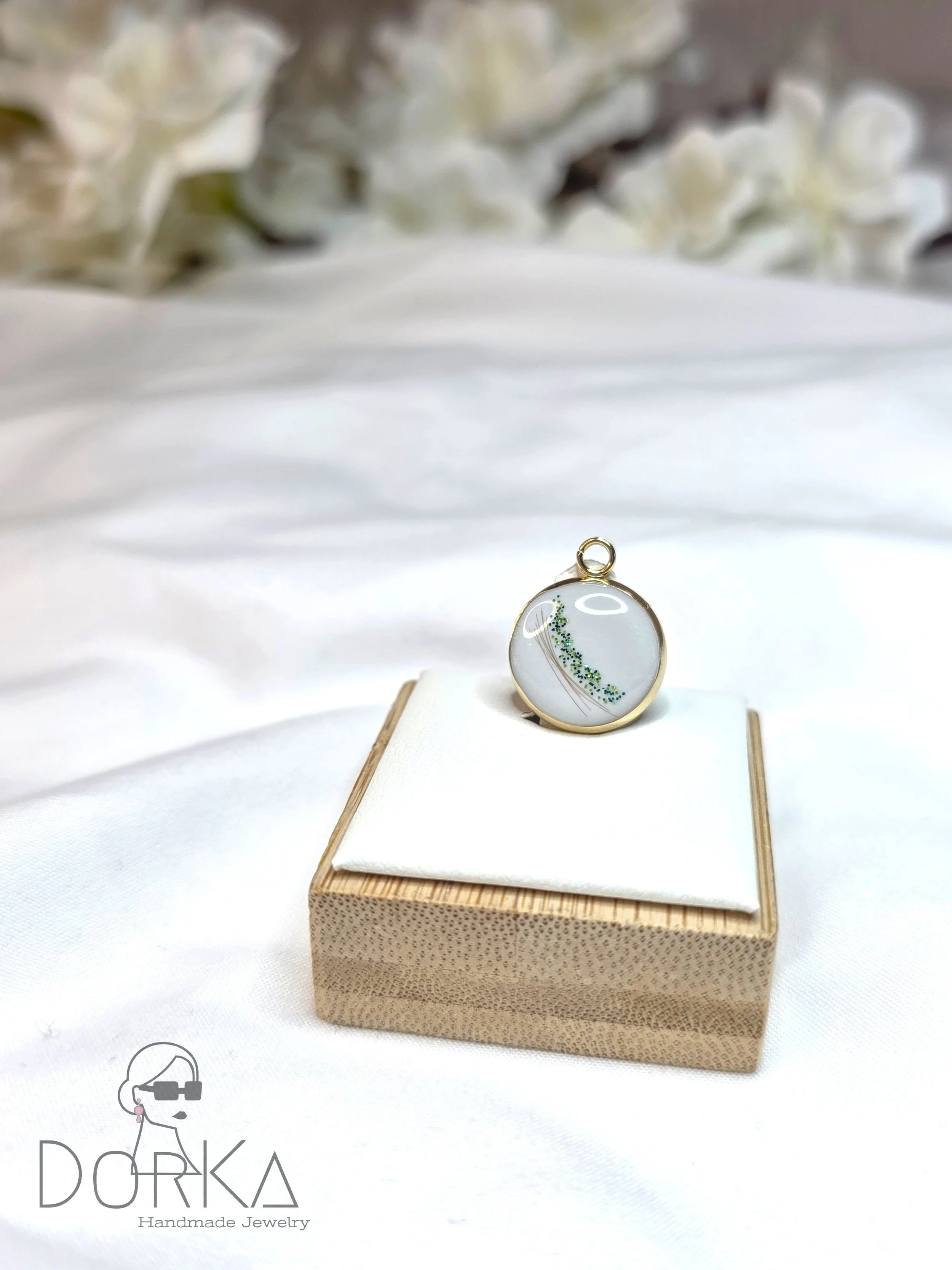 Goldring mit transparentem Anhänger mit grünen Dekorationen, auf einem weißen Kissen, auf einem kleinen Holzkästchen, Blumen im Hintergrund.