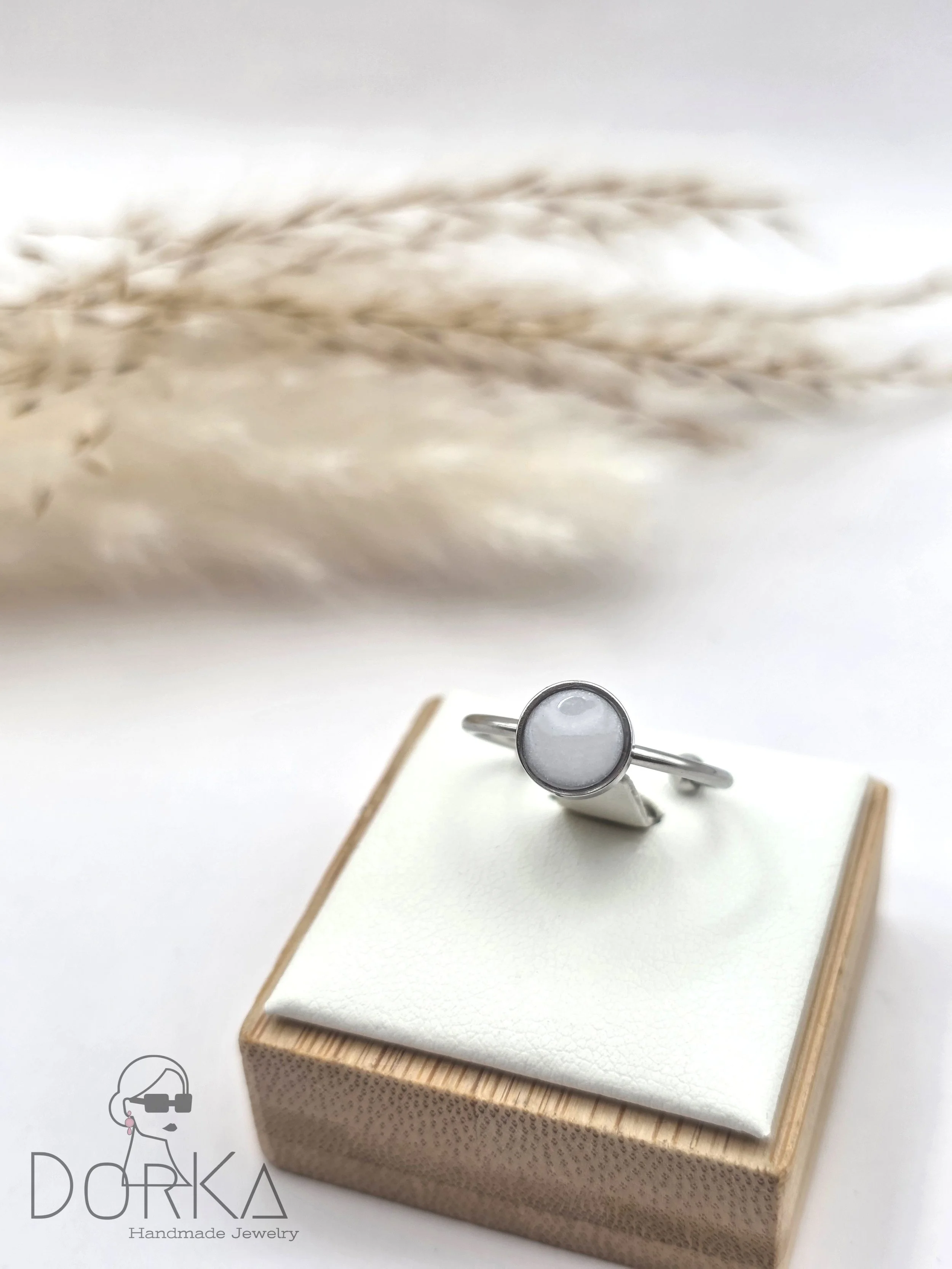 Silberner Ring mit grauem Stein auf einem weißen Schmuckkissen, Holzbasis, mit getrockneten Gräsern im Hintergrund.