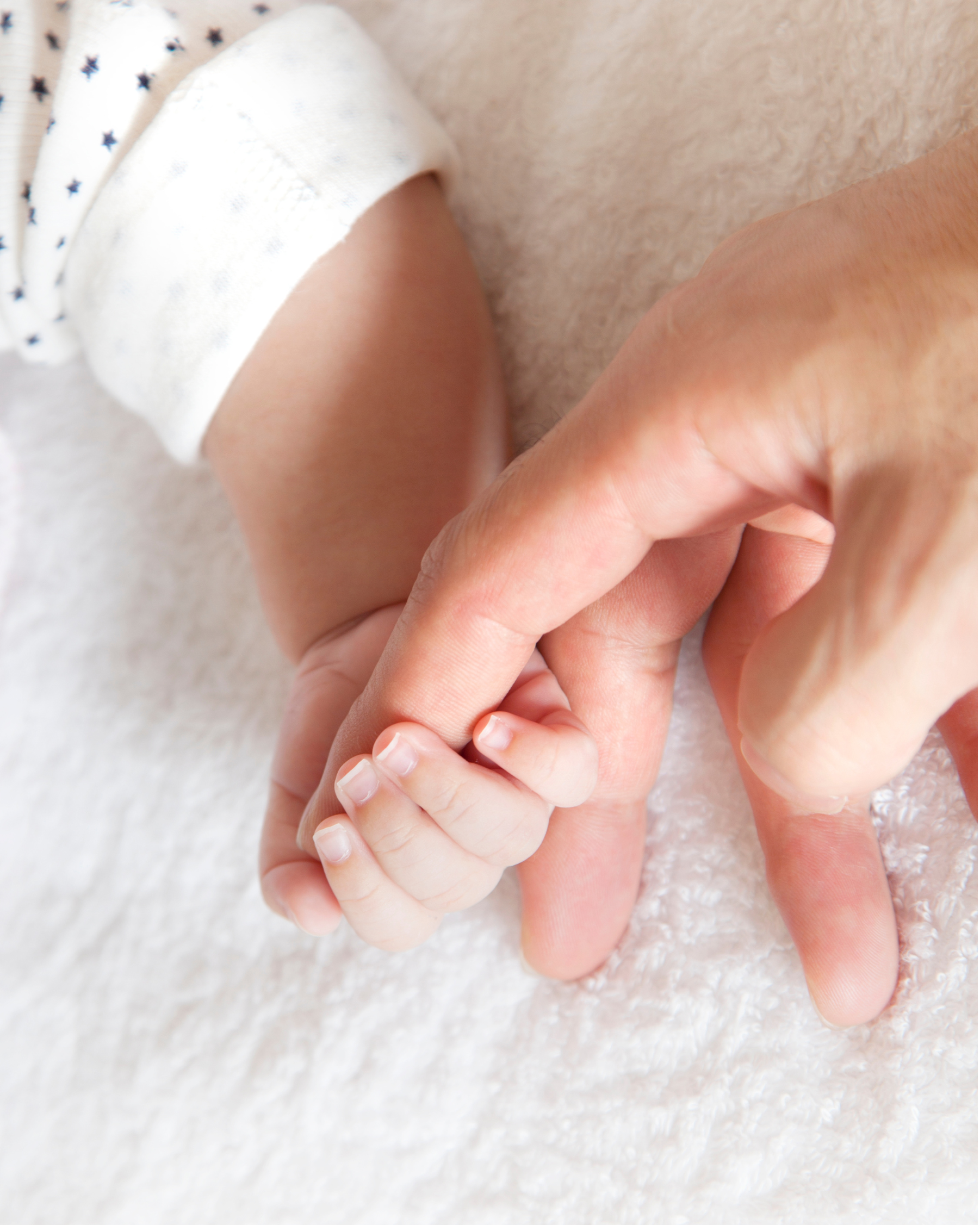 Nahaufnahme einer Erwachsenenhand, die einen kleinen Babyfinger hält, auf einem weichen, hellen Stoffuntergrund.