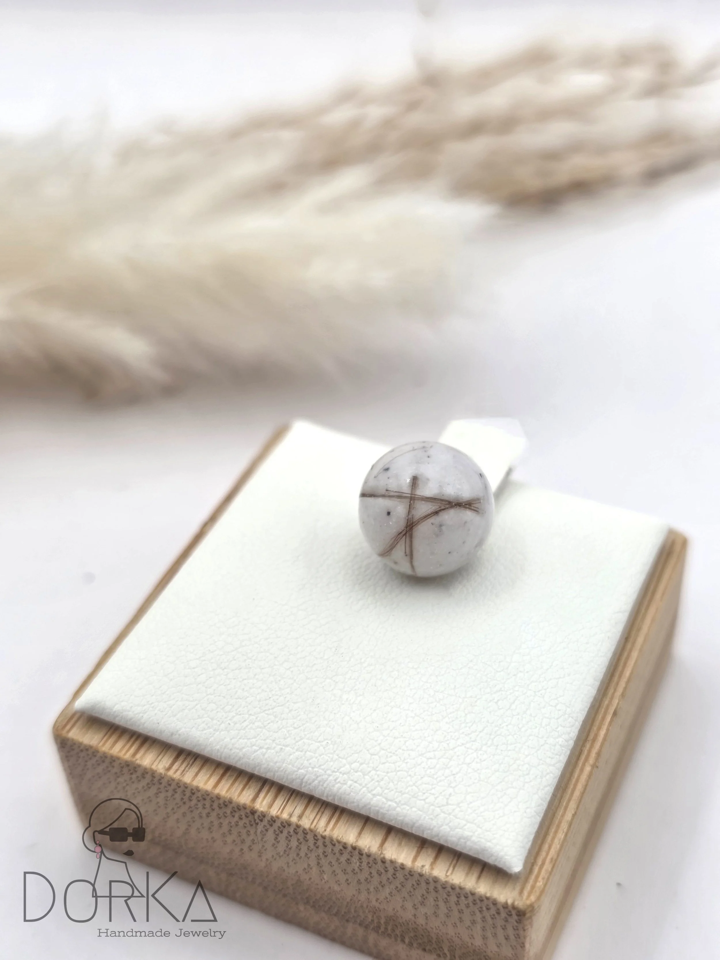 Handgefertigter Schmuck mit einer weißen Marmorkugel mit braunen Linien, präsentiert auf einem weißen Kissen in einer Holzschachtel.