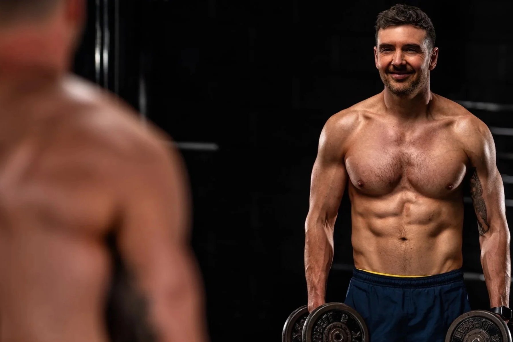 A shirtless man holding dumbbells and smiling, facing another shirtless man in a gym setting with a dark background.